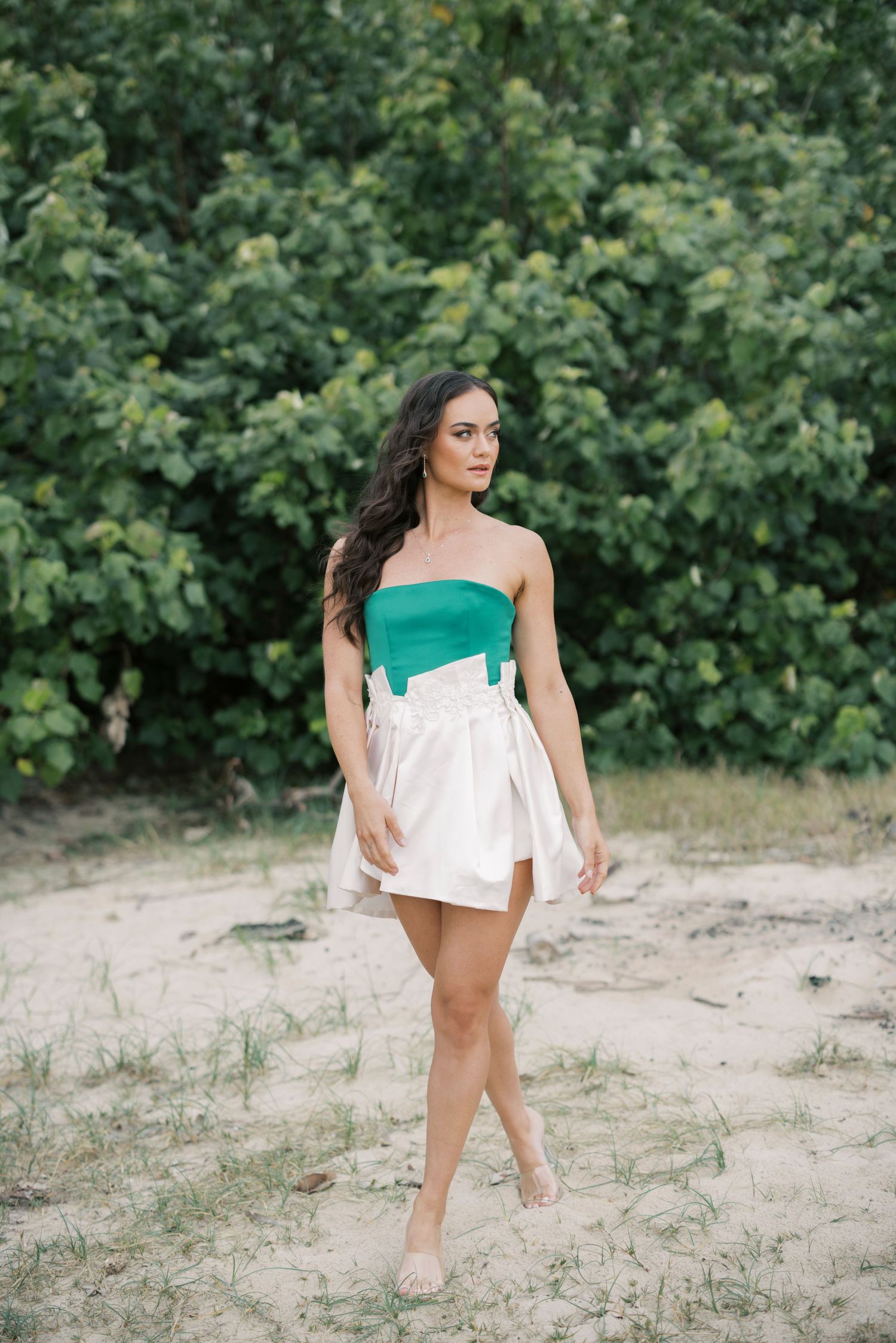 Woman in green and white dress poses gracefully against lush foliage background in outdoor photoshoot.