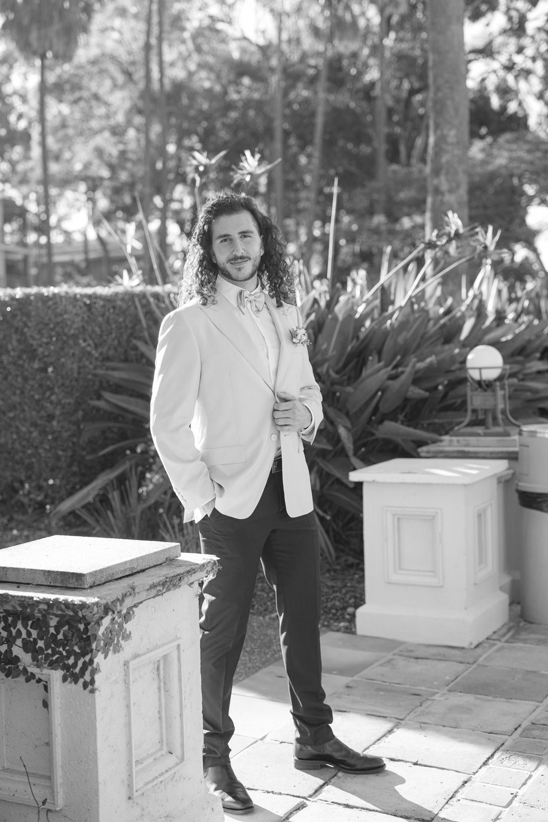 A person in a white top and dark pants sits on a stone ledge in a garden setting with plants in the background.