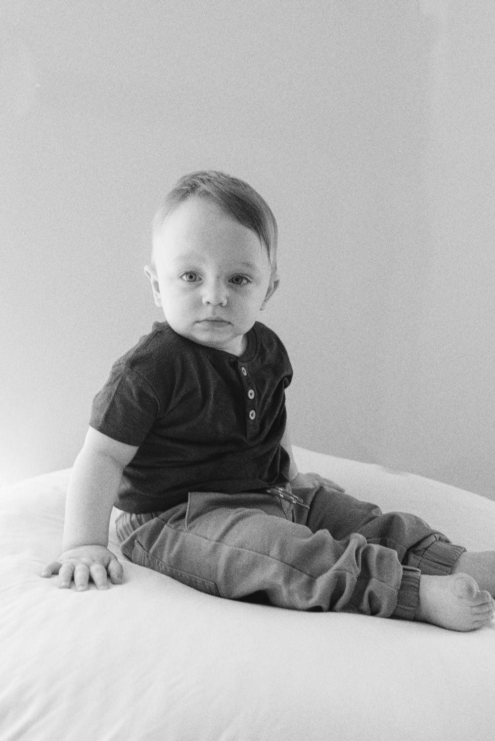 Baby in black polo shirt and gray pants sitting on white surface.