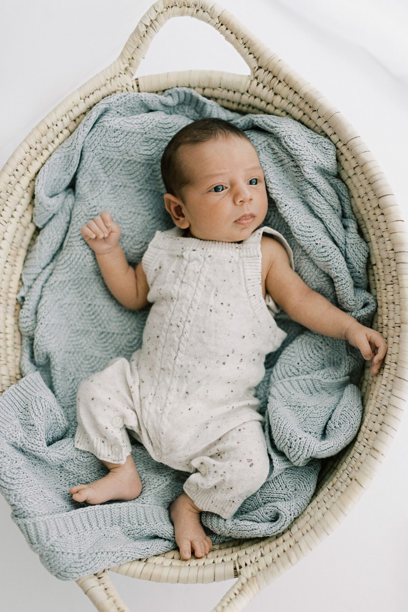 A baby in a white knit outfit lies in a round woven basket with a soft blue blanket.