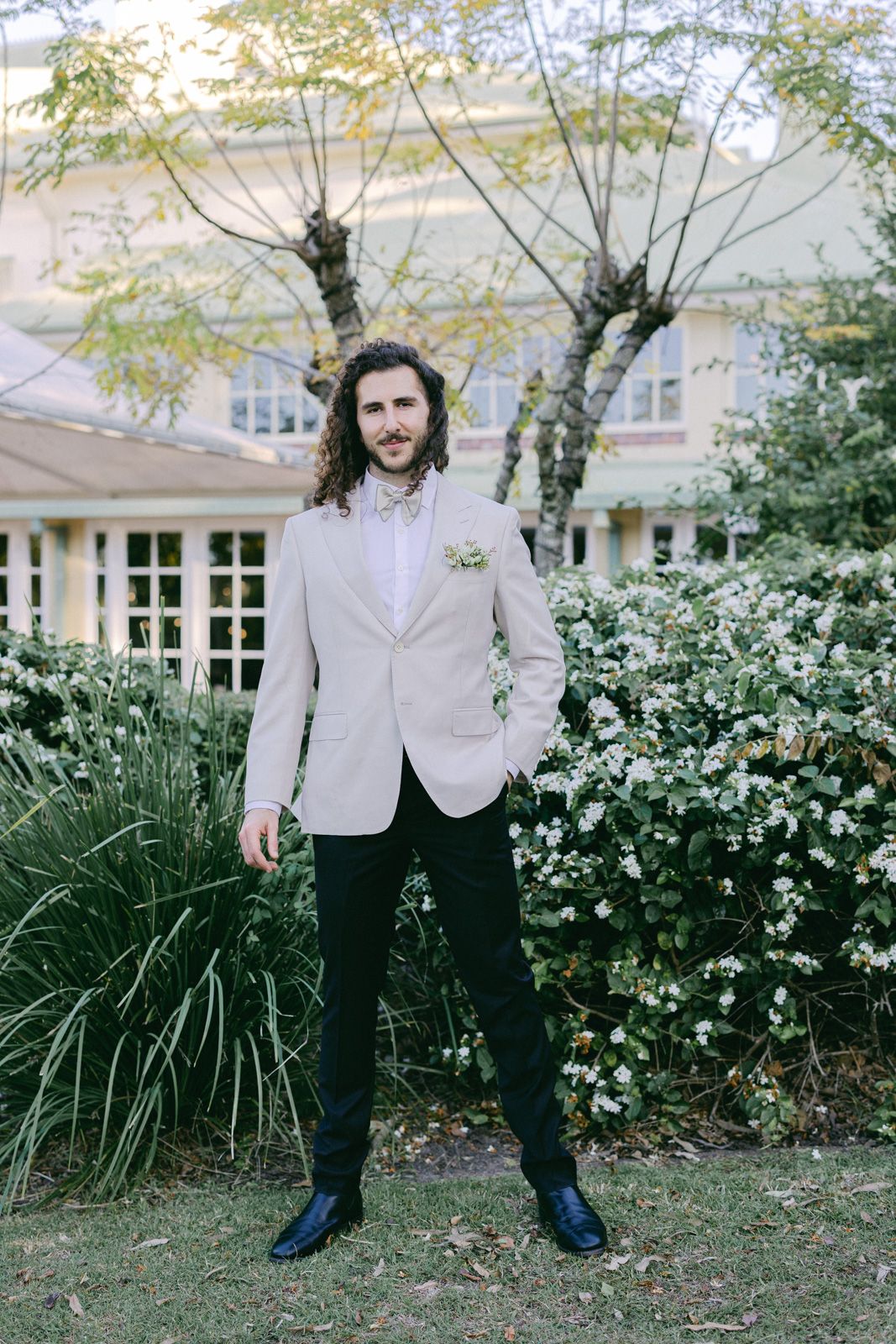 A figure in a light gray suit jacket and dark pants stands in front of lush garden landscaping.