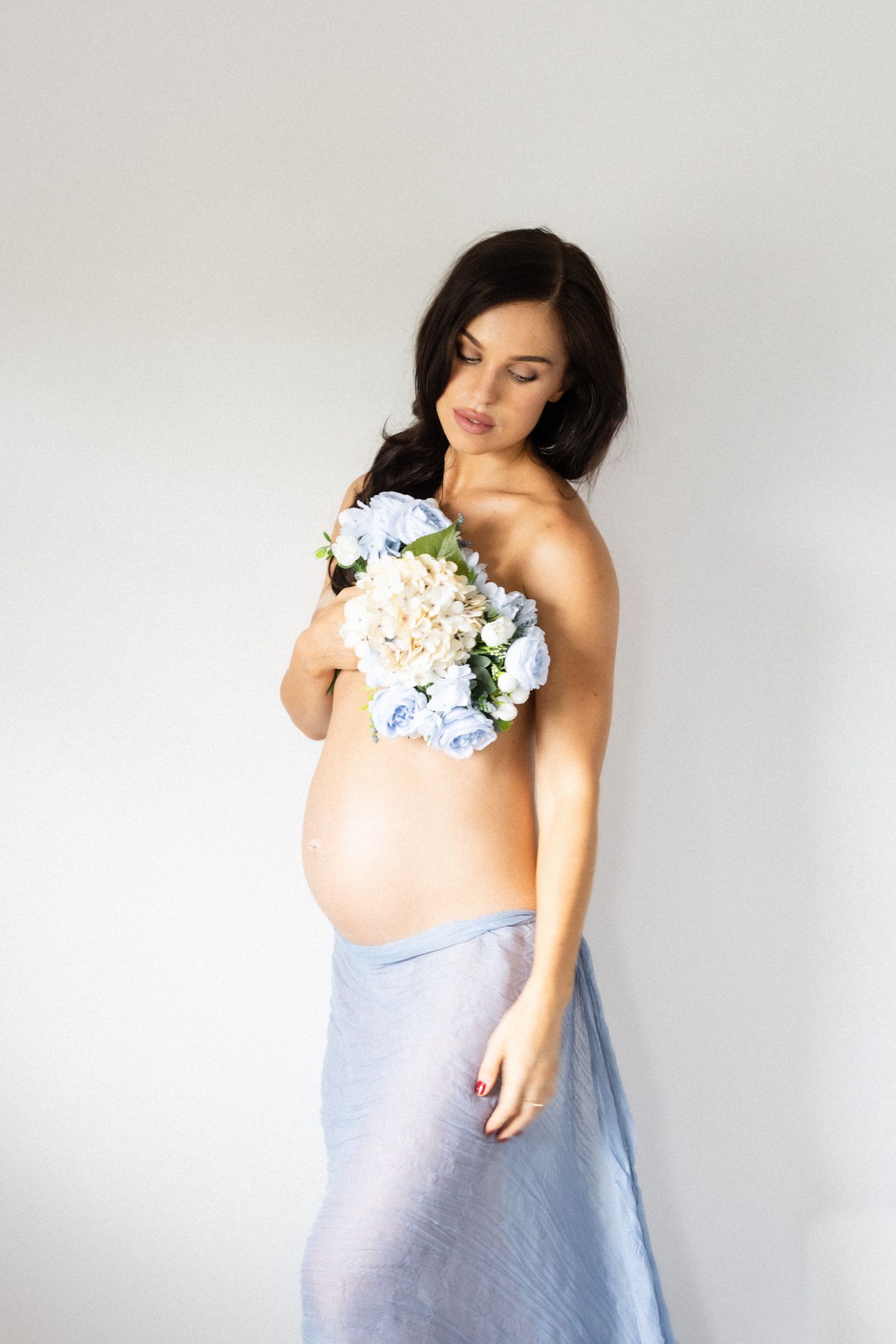 Ethereal maternity portrait with blue flowers and draped fabric against white background.