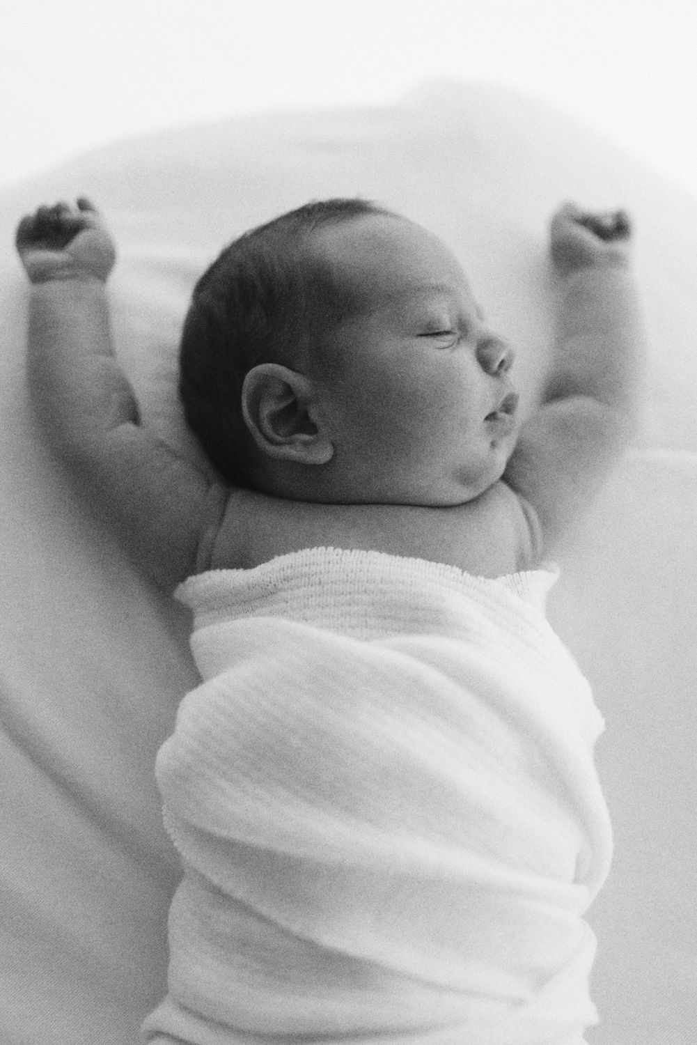 Black and white image of sleeping newborn with raised arms wearing white swaddle.