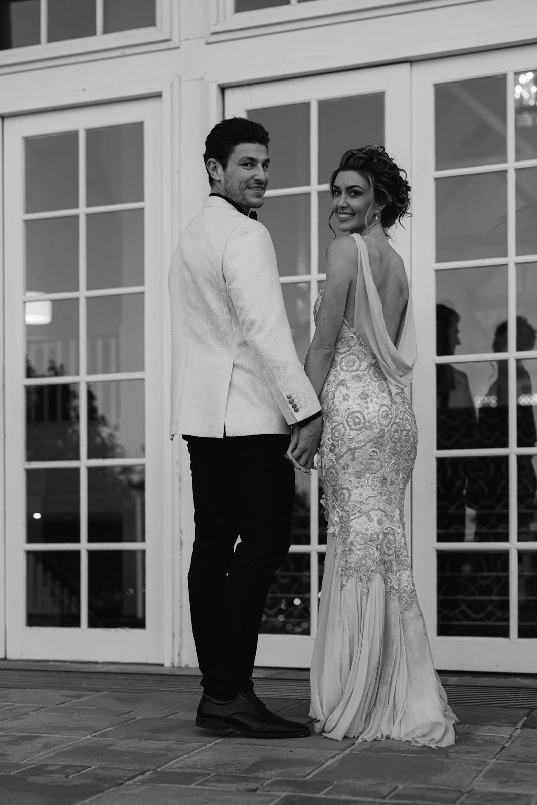 A black and white wedding photo showing a couple in formal attire posing together in front of glass doors.