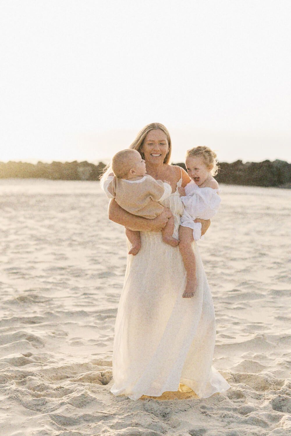 Smiling blonde mother in white maxi dress holding two young children on a golden sandy beach at sunset.