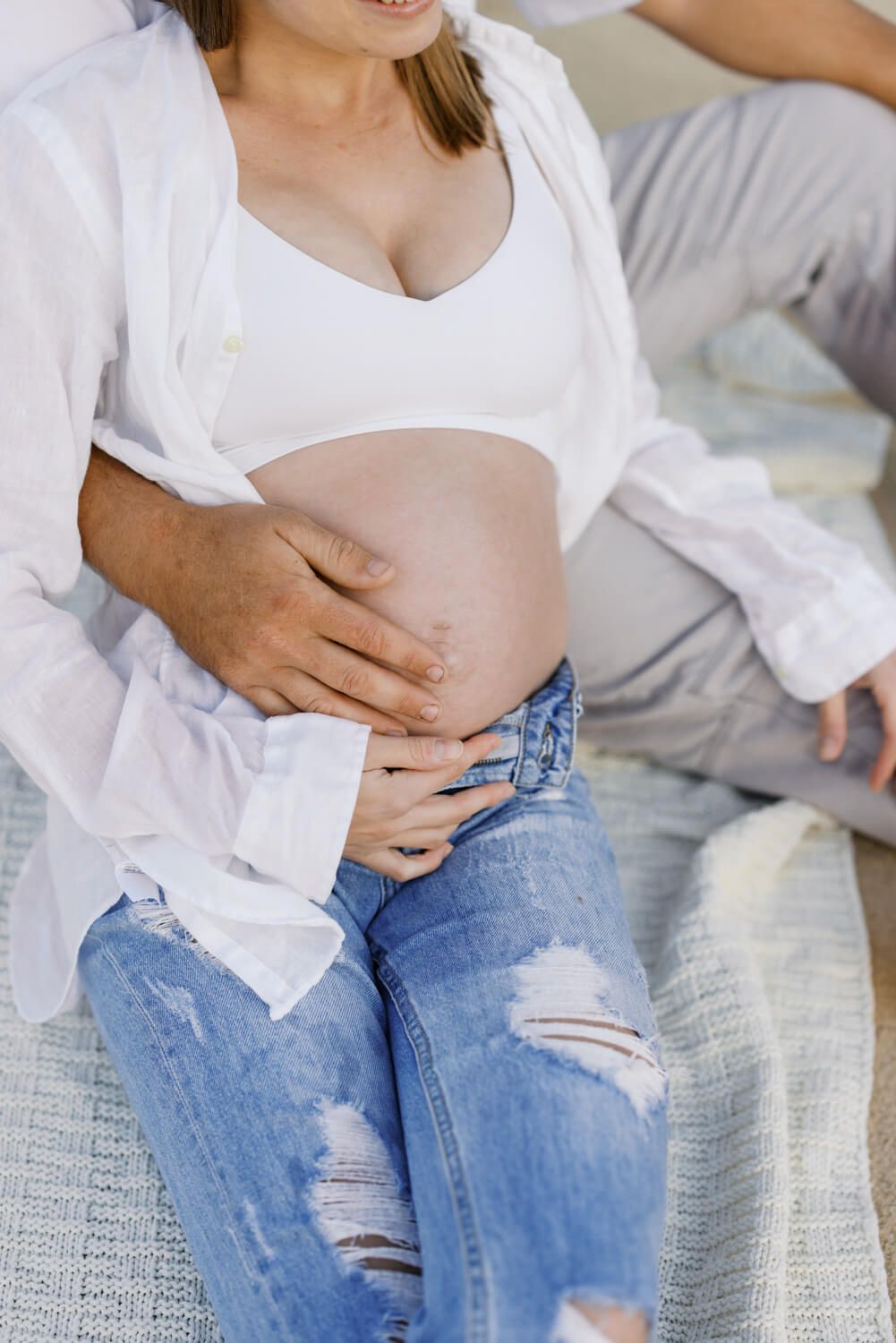 Pregnant woman in white bra and ripped jeans sitting on bed with partner's hands gently cradling her baby bump.