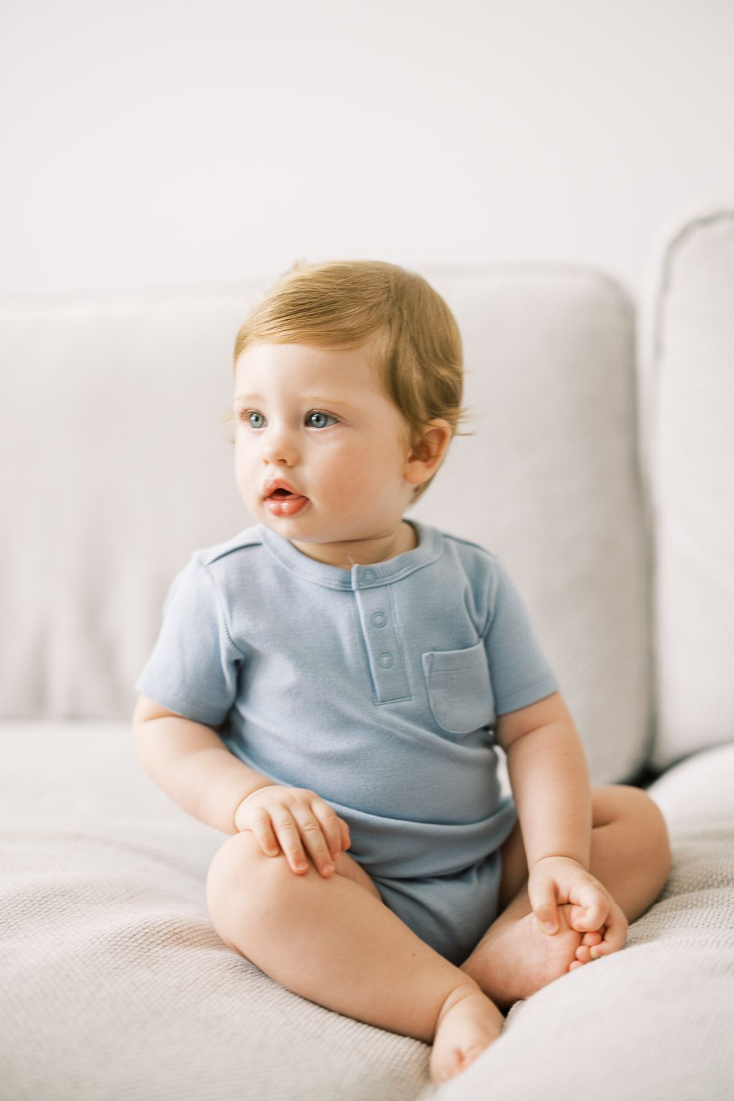 Baby in blue romper sitting on white couch looking to the side.