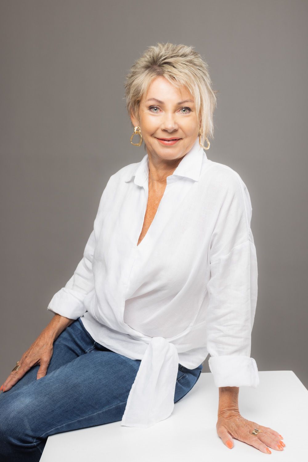 A person wearing a white button-down shirt and jeans poses confidently in a studio setting against a gray background.
