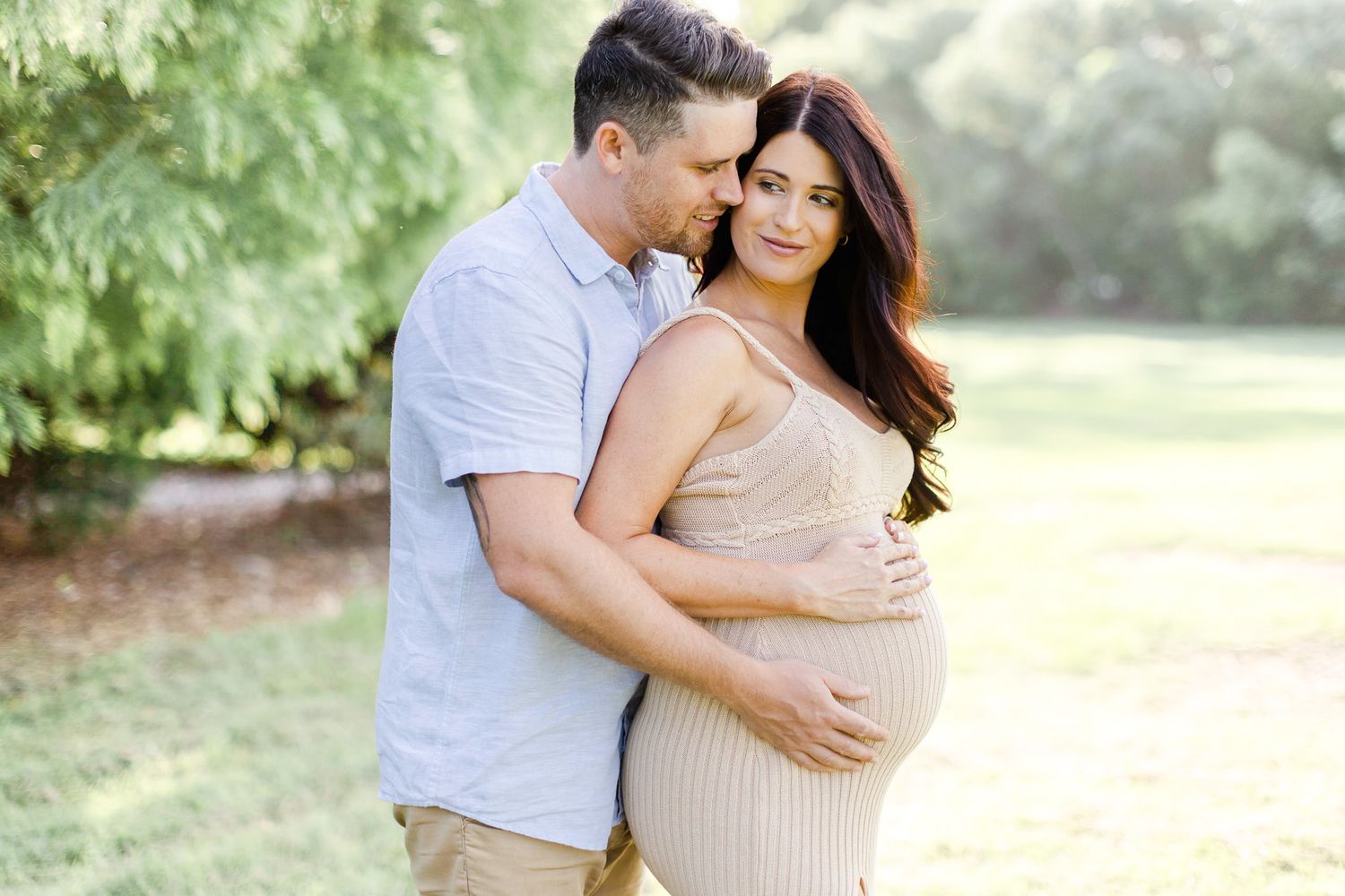 A couple shares tender moments in a sun-drenched outdoor setting wearing light neutral-colored clothing.