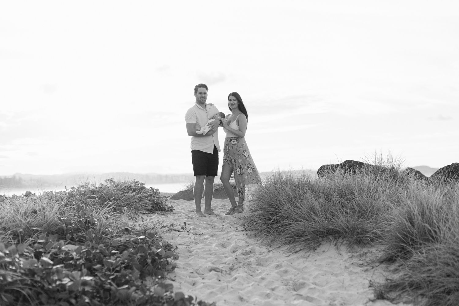 A couple stands together on a sandy coastal path with tall beach grass and ocean views in black and white.