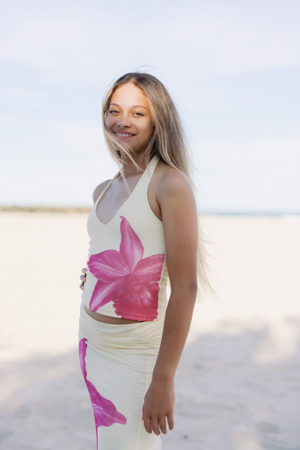Smiling young girl with long blonde hair wearing a floral crop top and skirt set on a sandy beach.