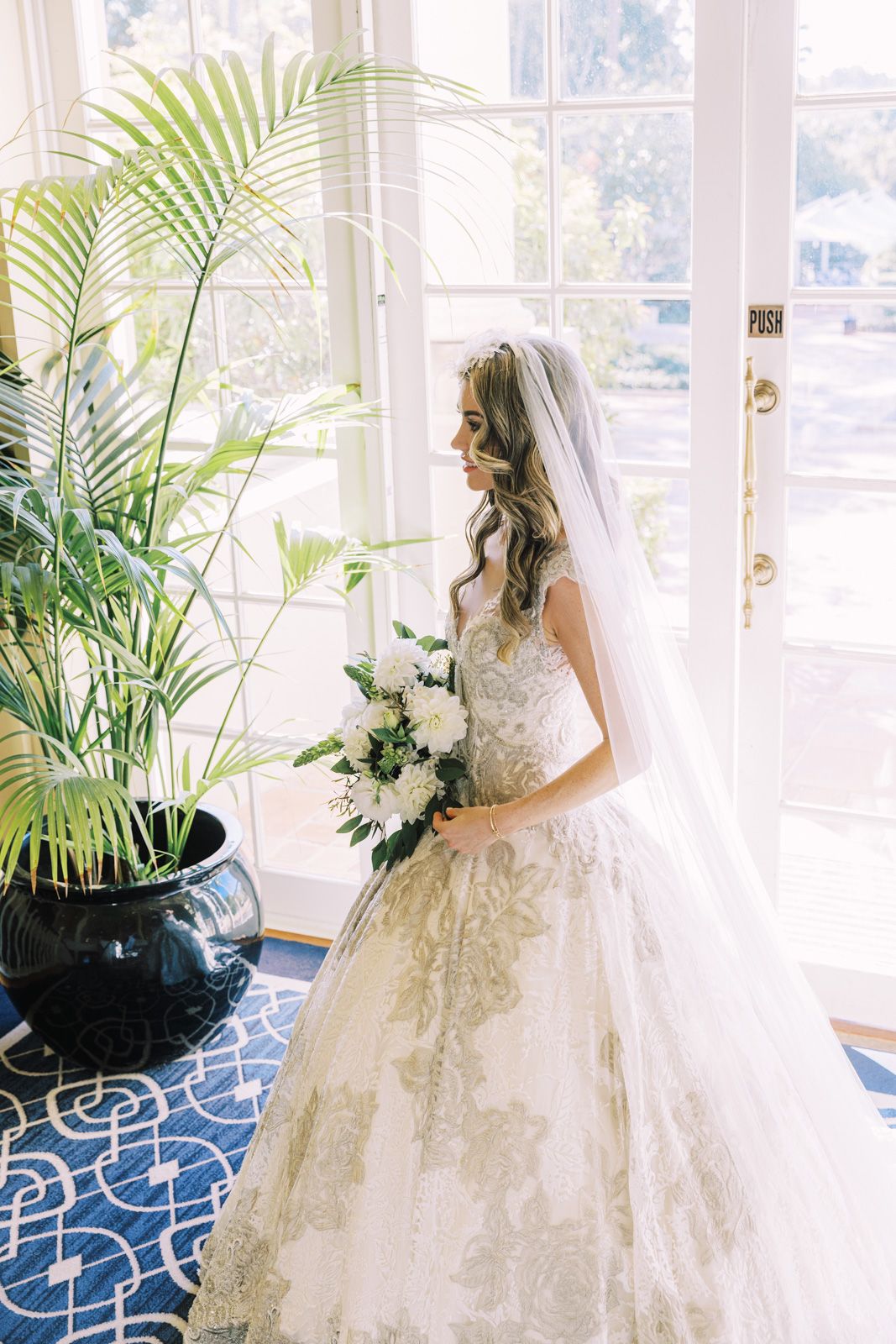 A bride in a luxurious white lace ball gown poses by bright windows with palm plants and white floral bouquet.