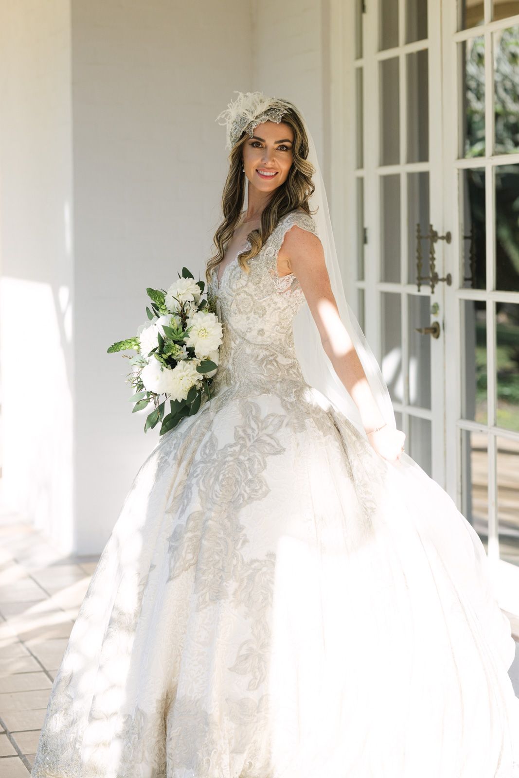 A bride in a flowing white lace wedding dress poses gracefully on a bright sunlit porch with French doors.