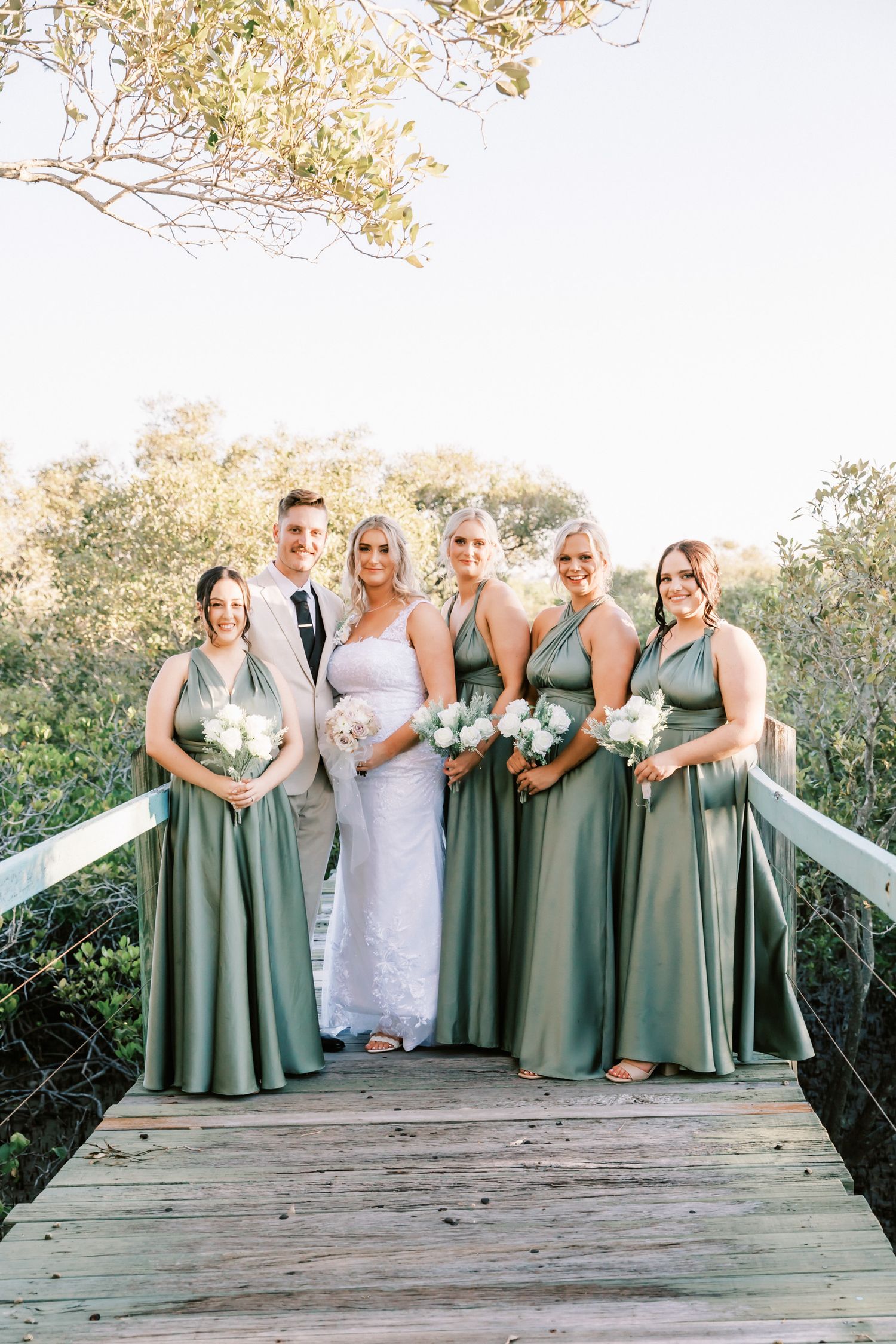 Wedding party in sage green bridesmaid dresses poses together on a wooden deck outdoors.