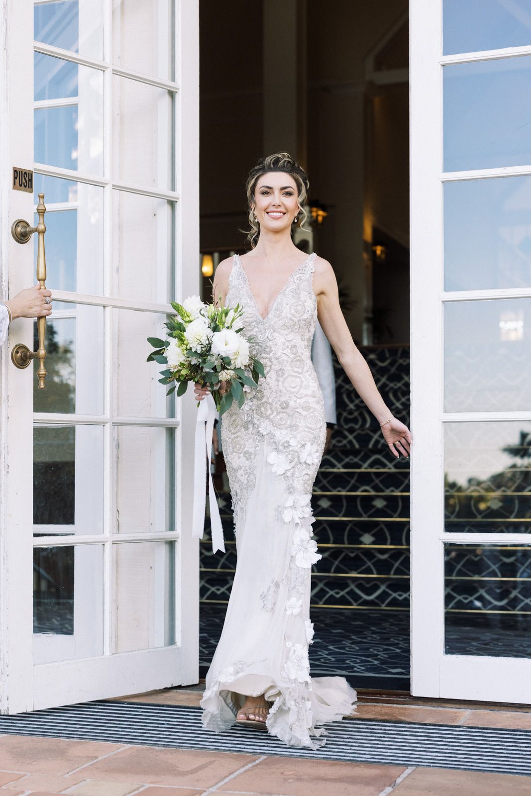 A bride in a fitted lace wedding gown with a plunging neckline stands in a doorway holding a white floral bouquet.