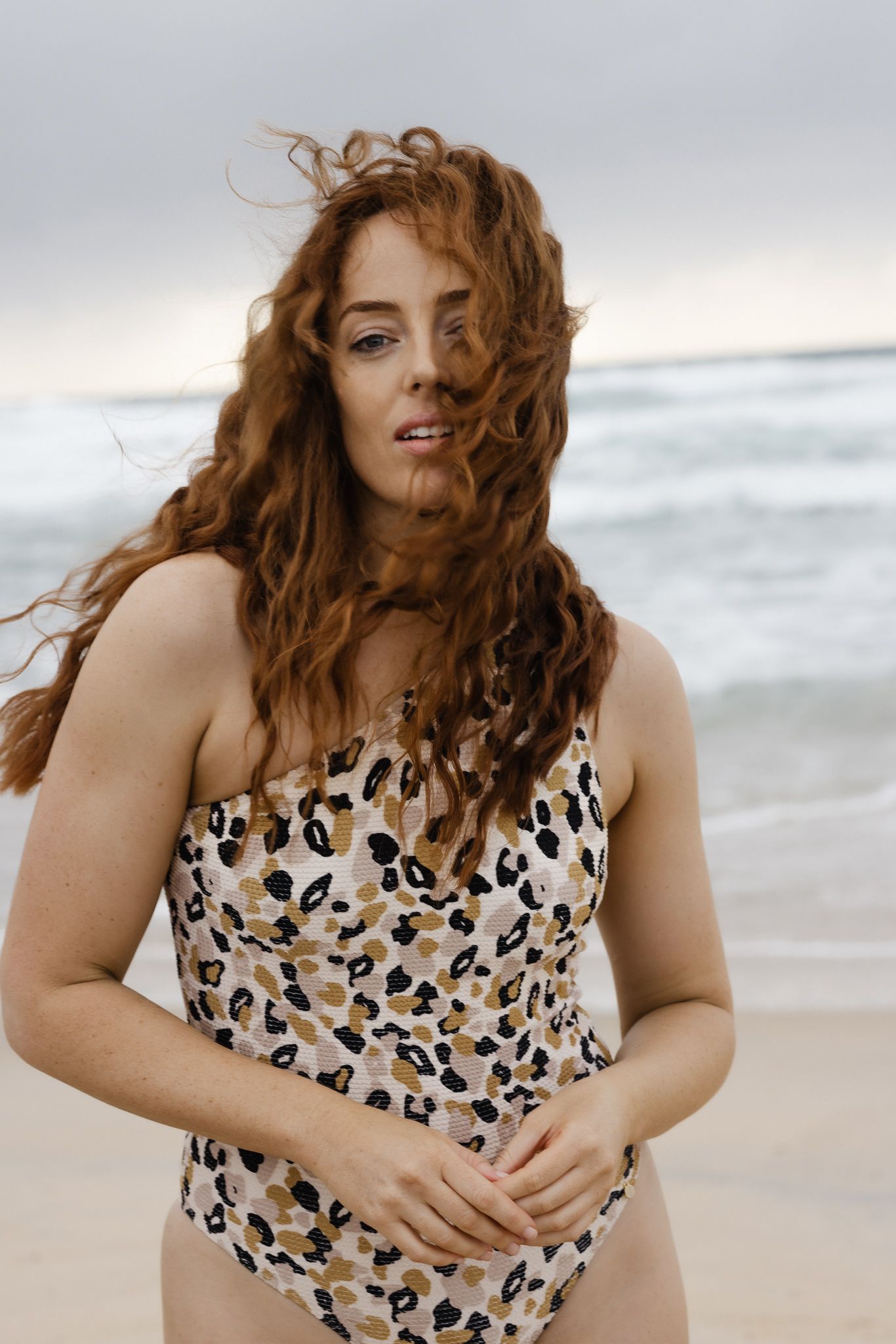 A person with windswept red curly hair poses on a beach wearing a leopard print swimsuit against an overcast sky.