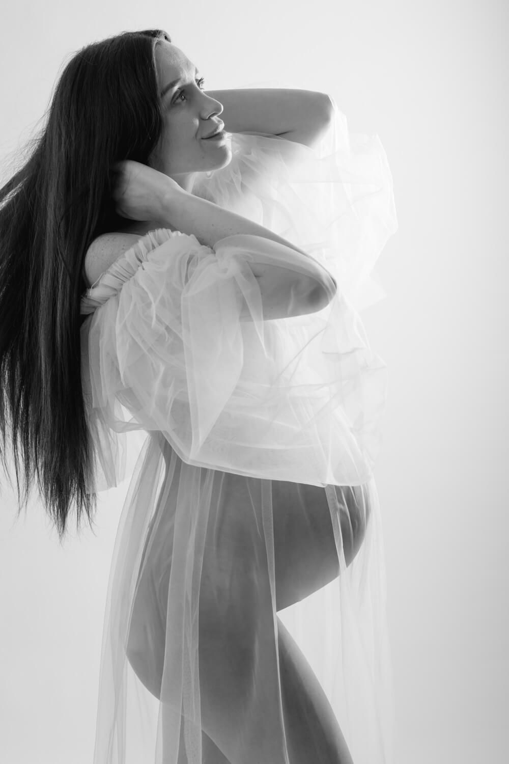 Black and white maternity photo of a smiling pregnant woman draped in sheer white fabric against a light background.