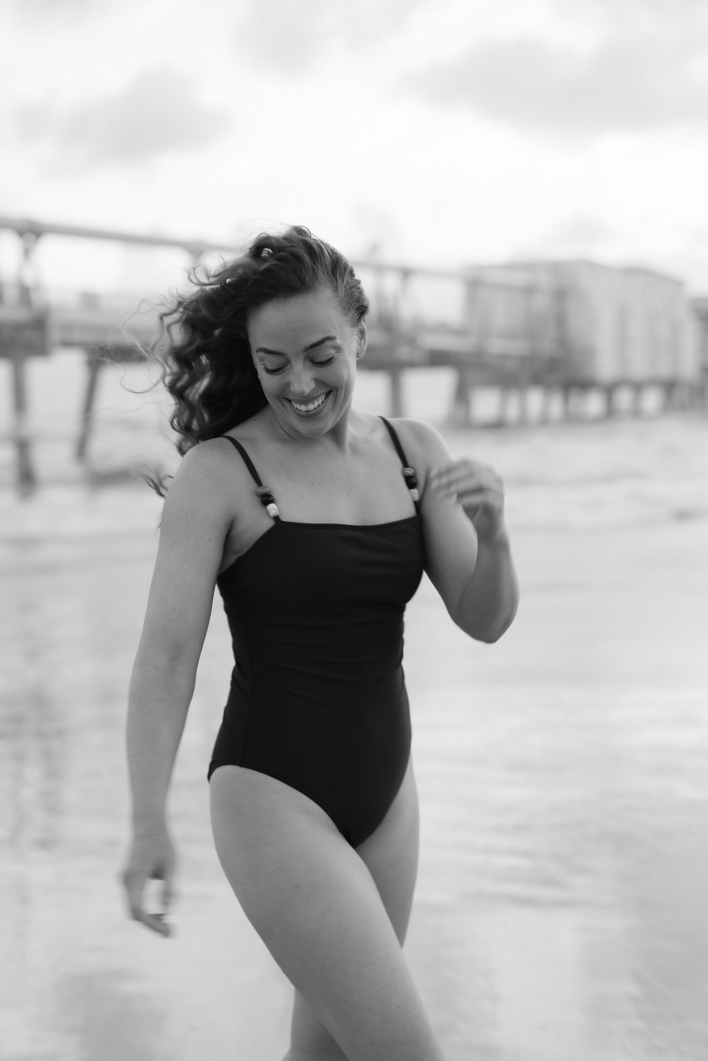 Smiling woman in black one-piece swimsuit on beach with pier in background, black and white photo.