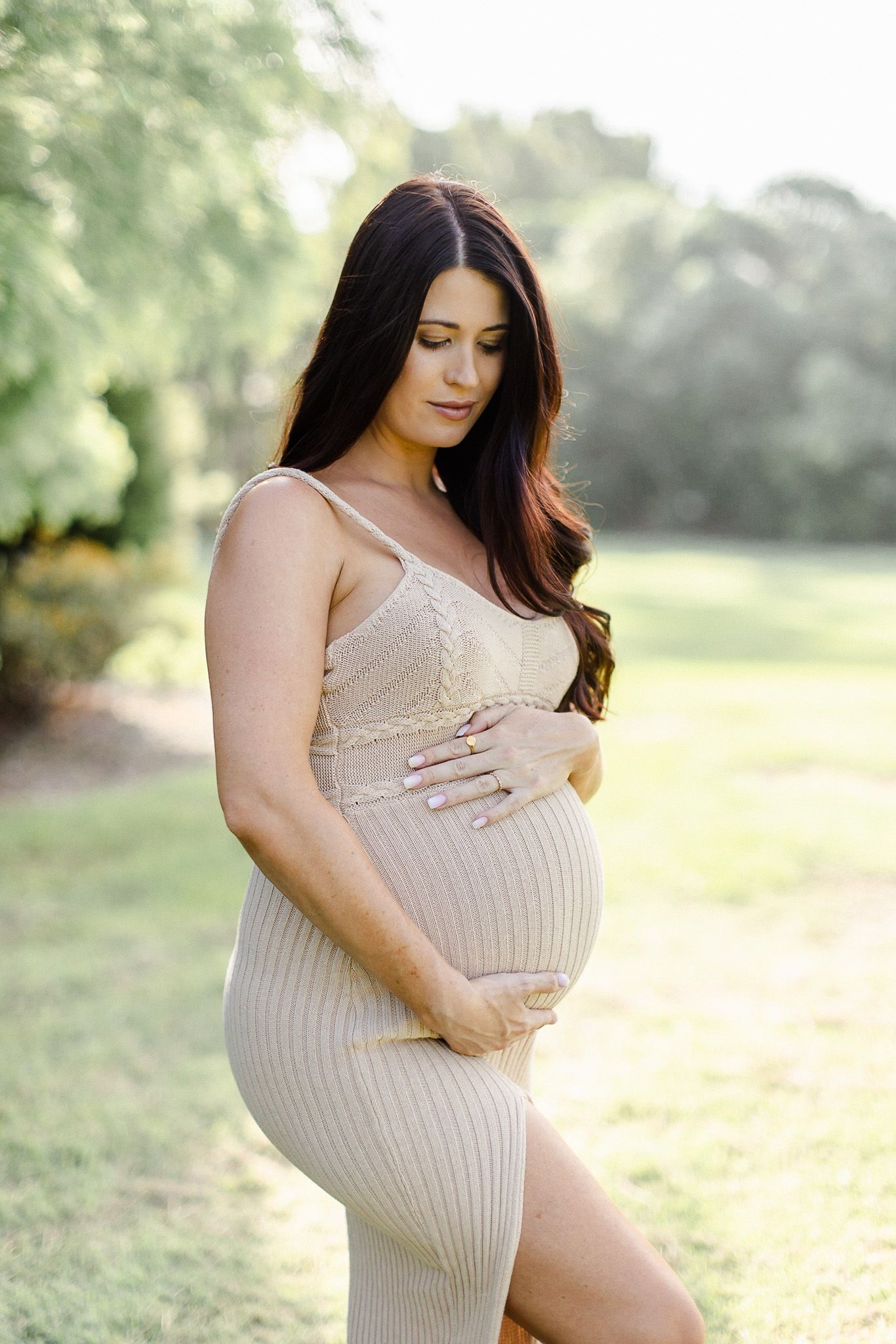 A maternity photoshoot in a park shows a woman in a beige knit dress posing against a soft natural backdrop.