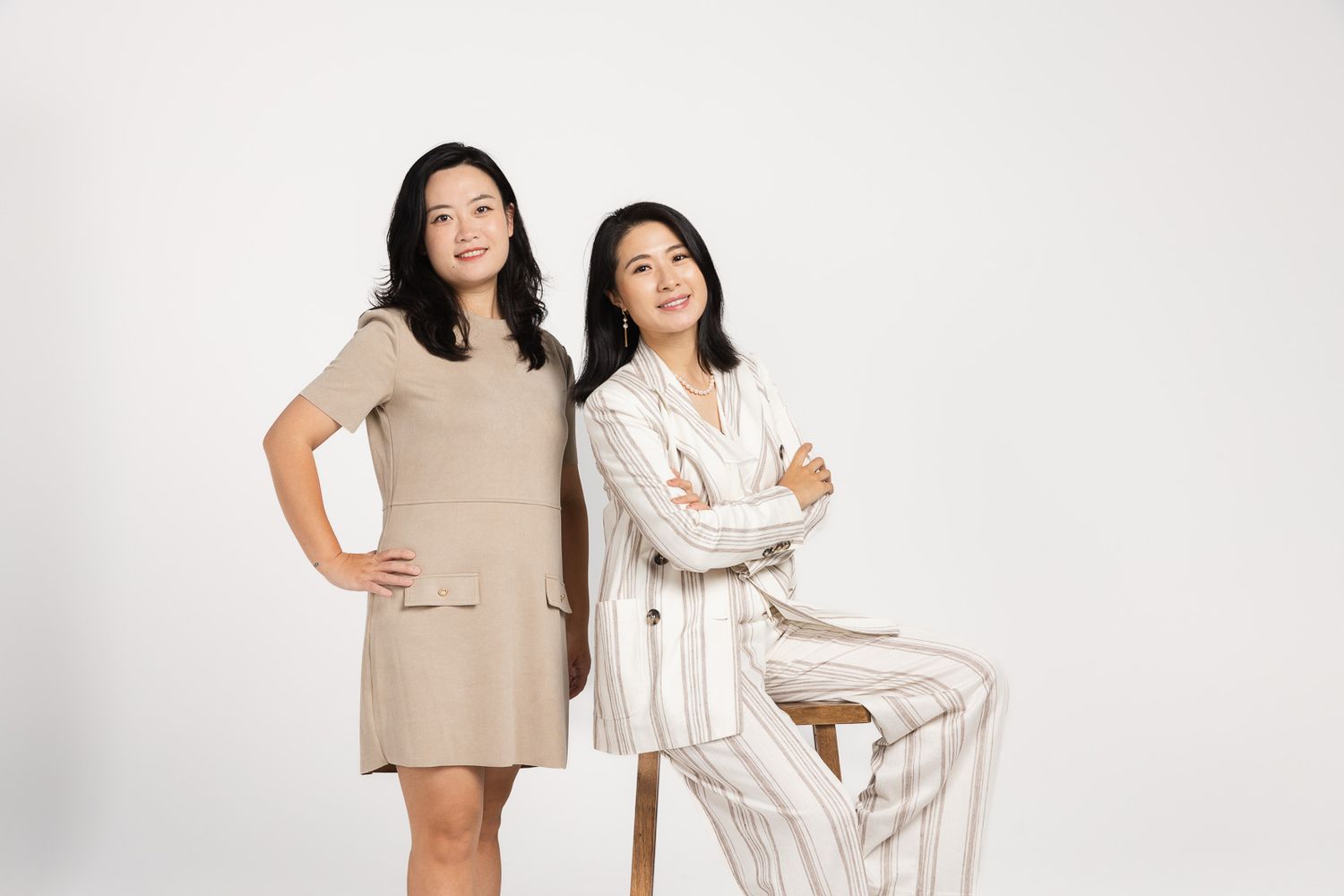 Two people pose together wearing coordinated beige and white outfits in a minimalist studio setting.