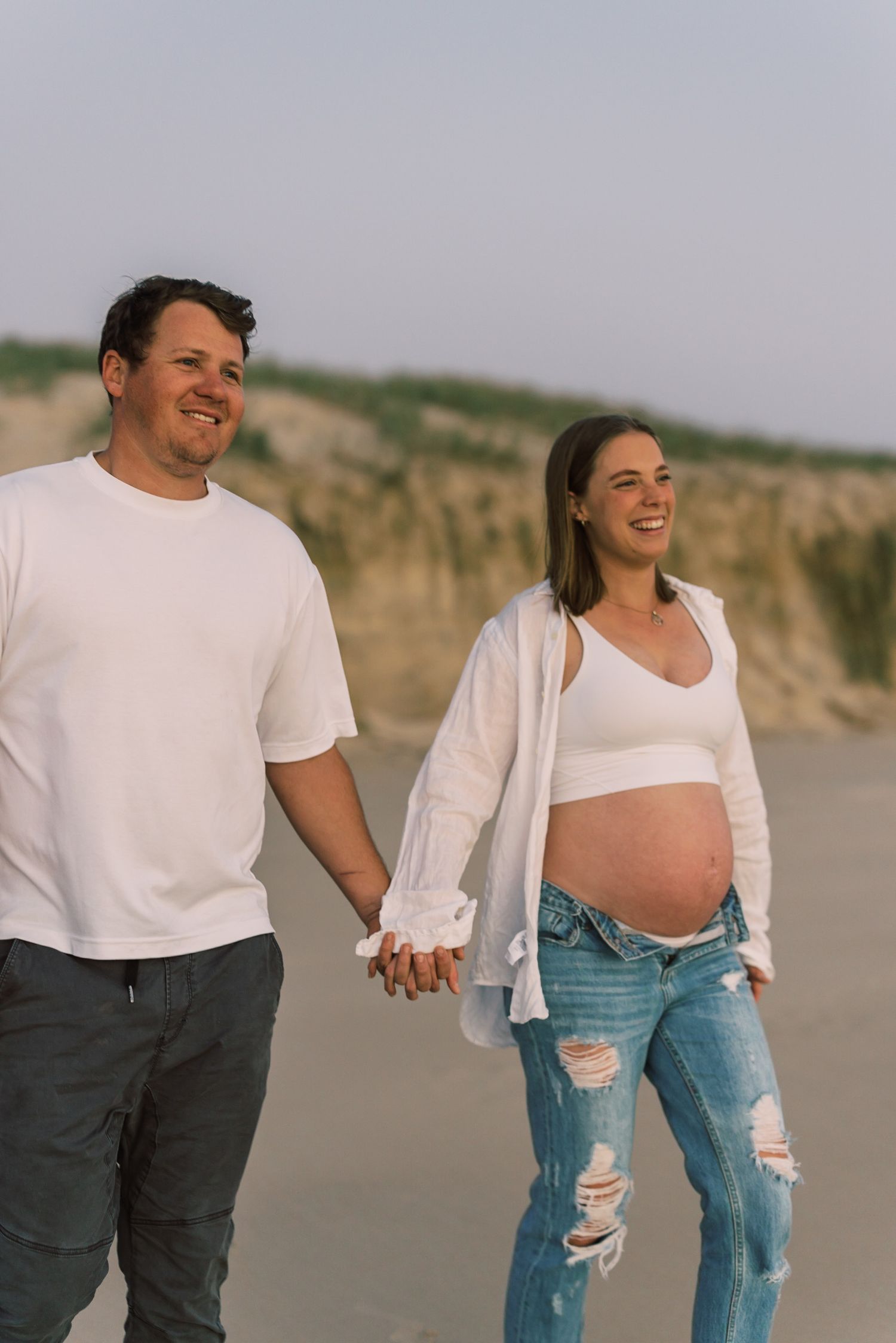 A casual couple walks hand in hand along a coastal path during a maternity photoshoot.