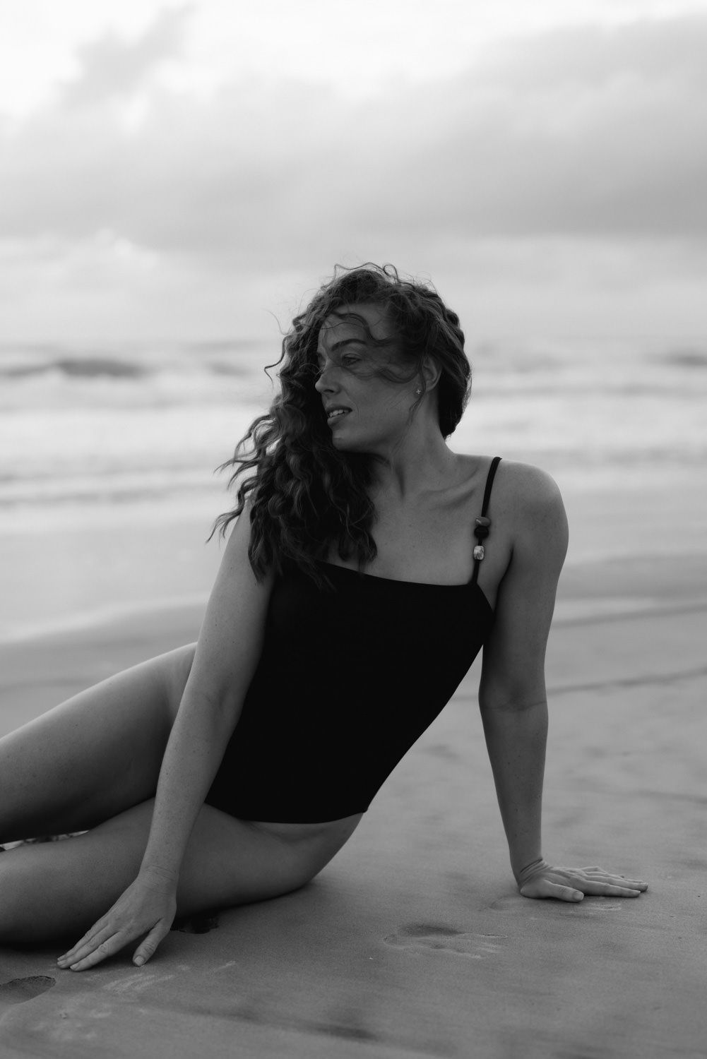 Black and white photo of a woman in a swimsuit sitting on a sandy beach with ocean waves in the background.