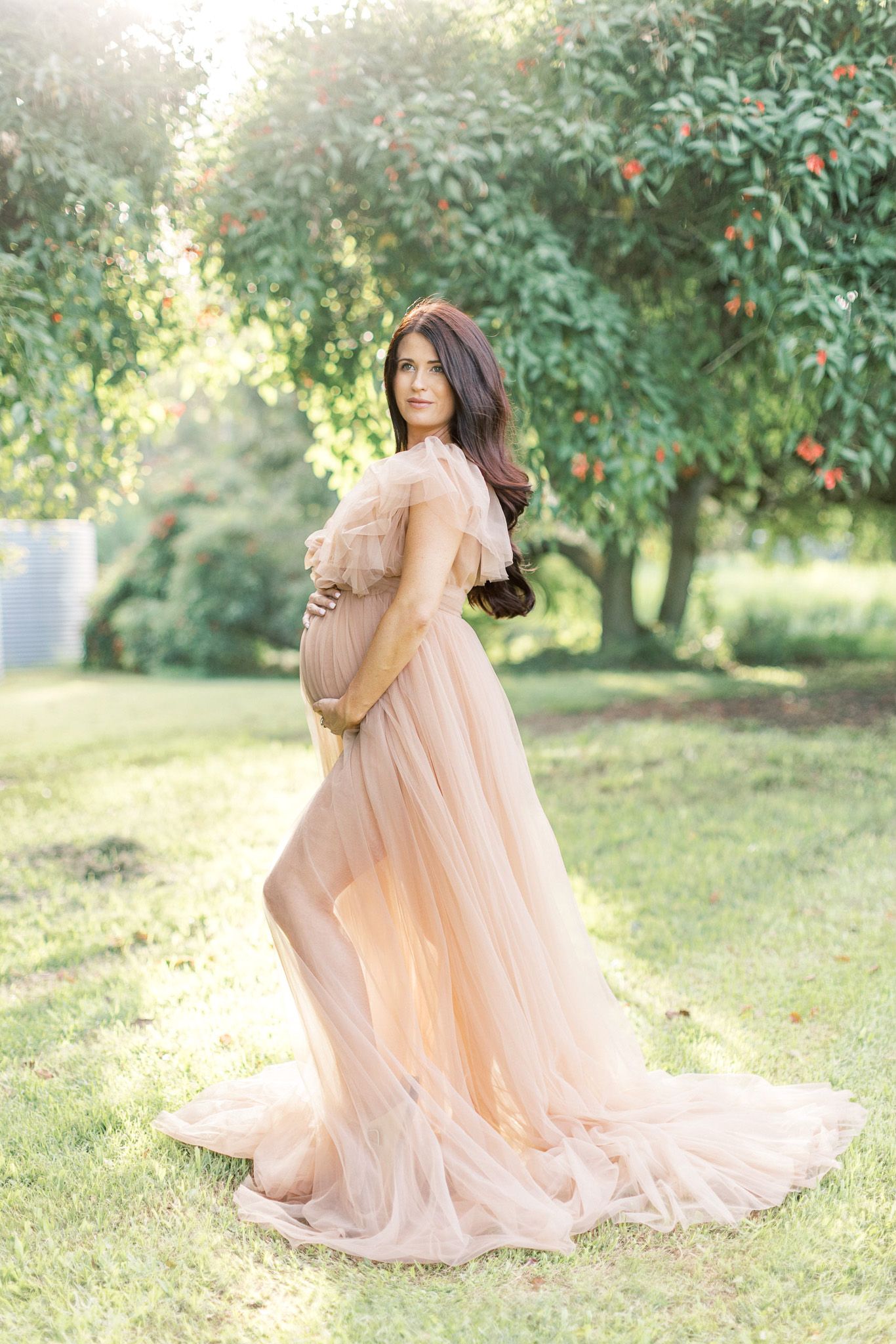 A flowing peach-colored tulle gown twirls gracefully in a sunlit garden setting with green trees in the background.