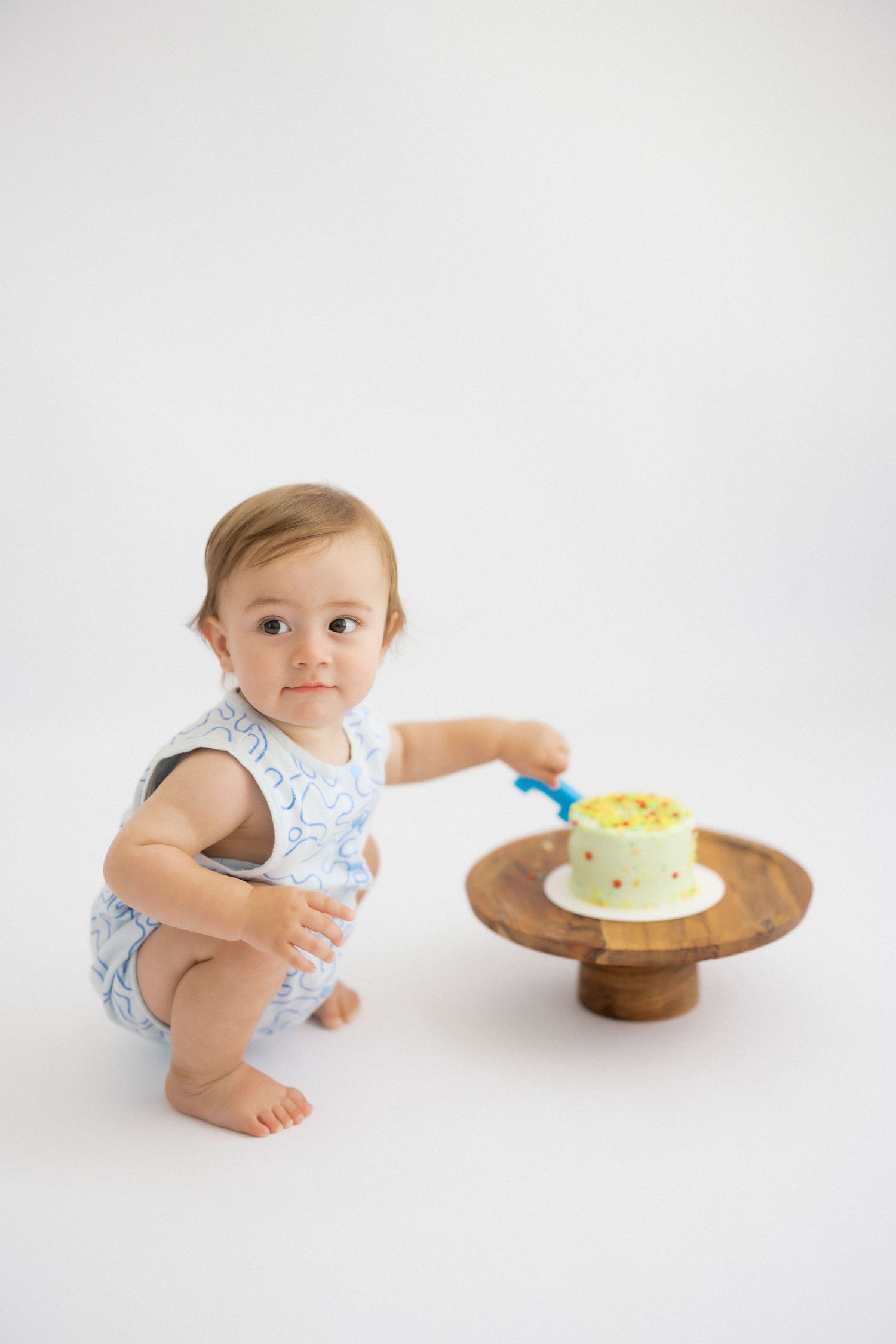 A baby in a floral romper curiously examines a small yellow cake on a wooden pedestal stand.