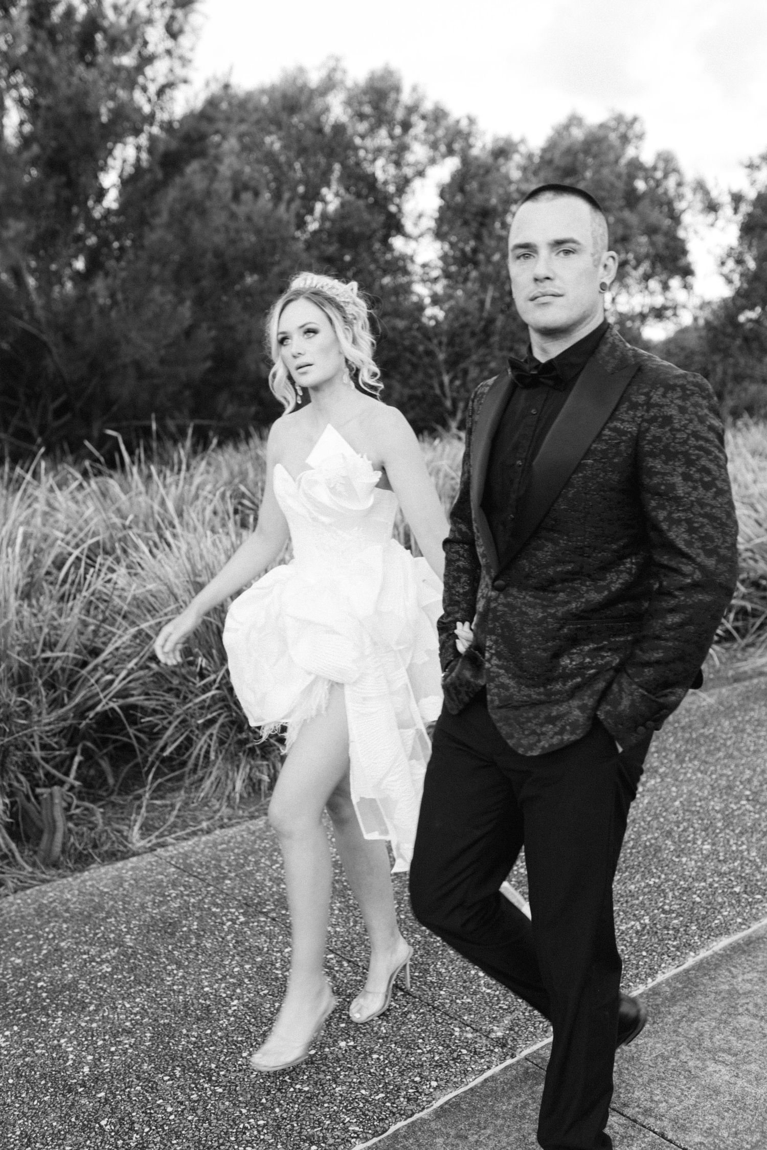 A formal couple poses on a pathway with natural foliage in black and white.