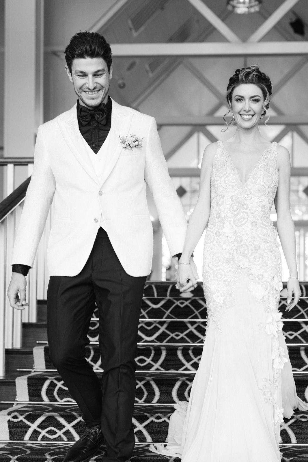 Black and white photo of newlyweds walking down ornate stairs in elegant wedding attire.