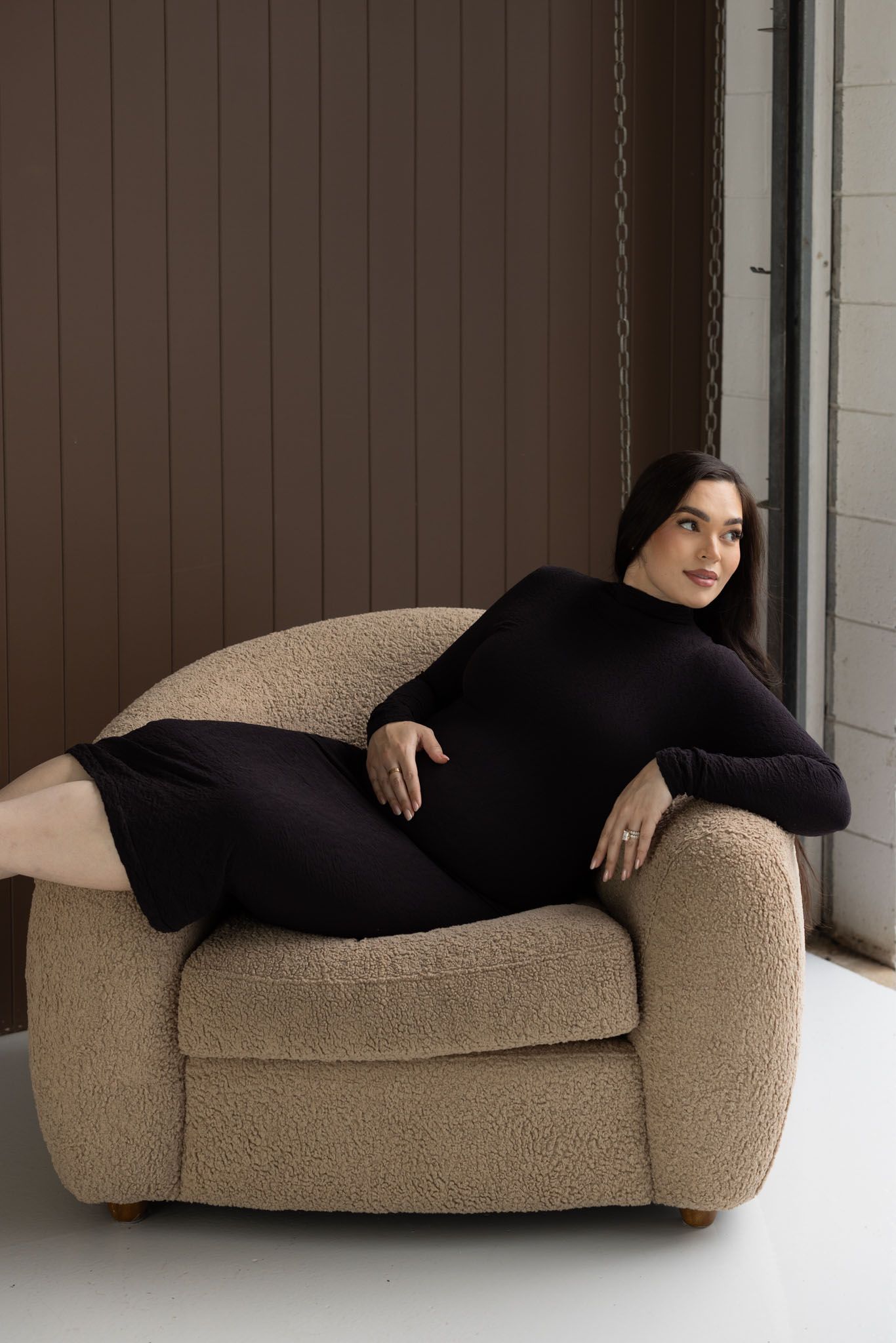 Pregnant woman studio photoshoot Gold Coast soft lightA woman in a black dress lounges elegantly on a plush beige armchair near a window.