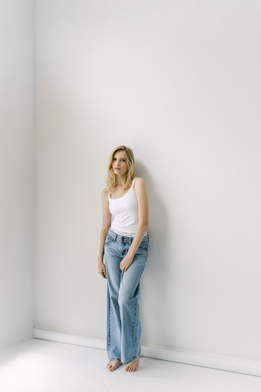 Blonde woman in white tank top and light wash flare jeans leaning against white wall.