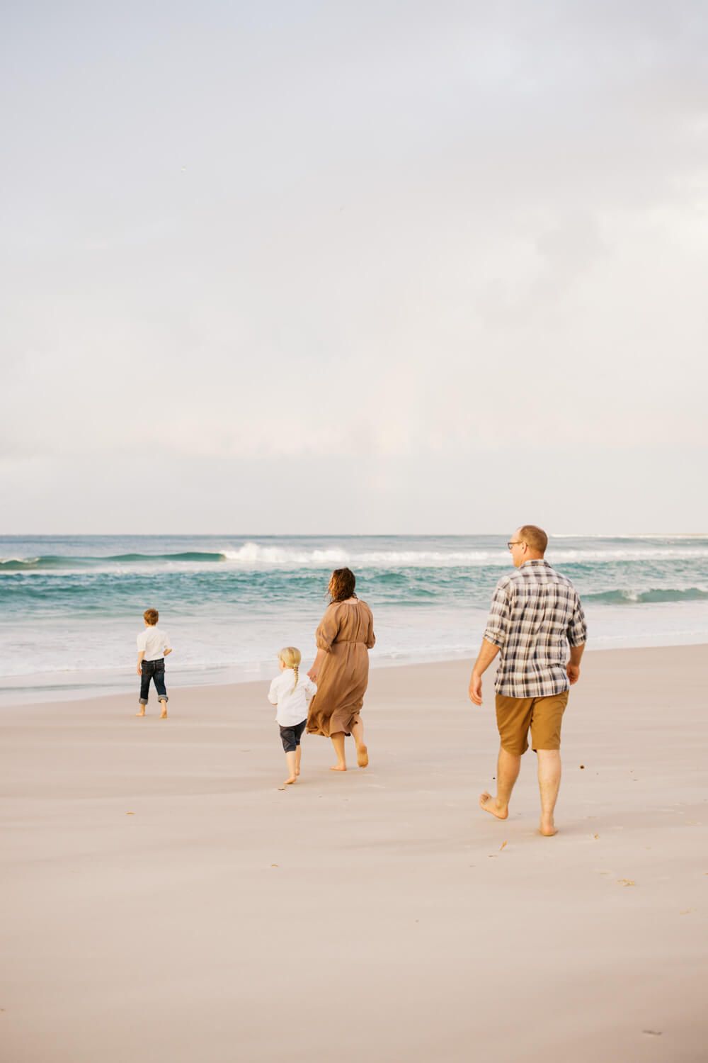 Family of four walking barefoot on a sandy beach toward ocean waves on a cloudy day.