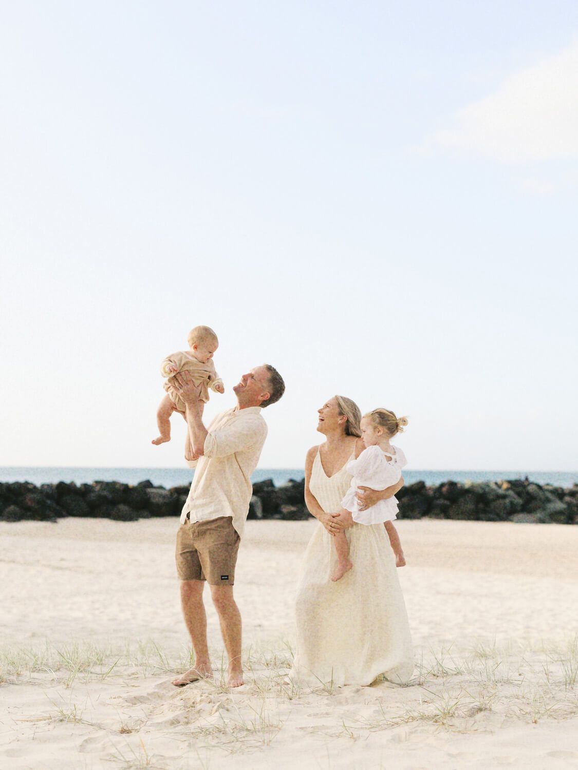 Family of four enjoying a beach day, dad lifting baby while mom holds toddler, wearing neutral tones on sandy shore.