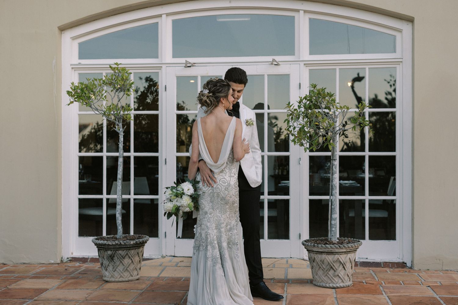 A couple embraces in front of elegant French doors with a backless beaded wedding gown and formal black attire.
