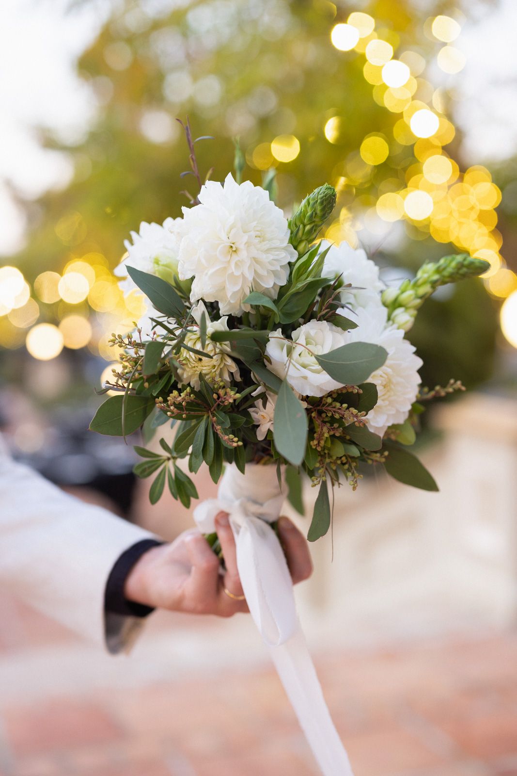 A white floral bouquet with eucalyptus leaves and ribbon is held against a soft bokeh background of golden lights.