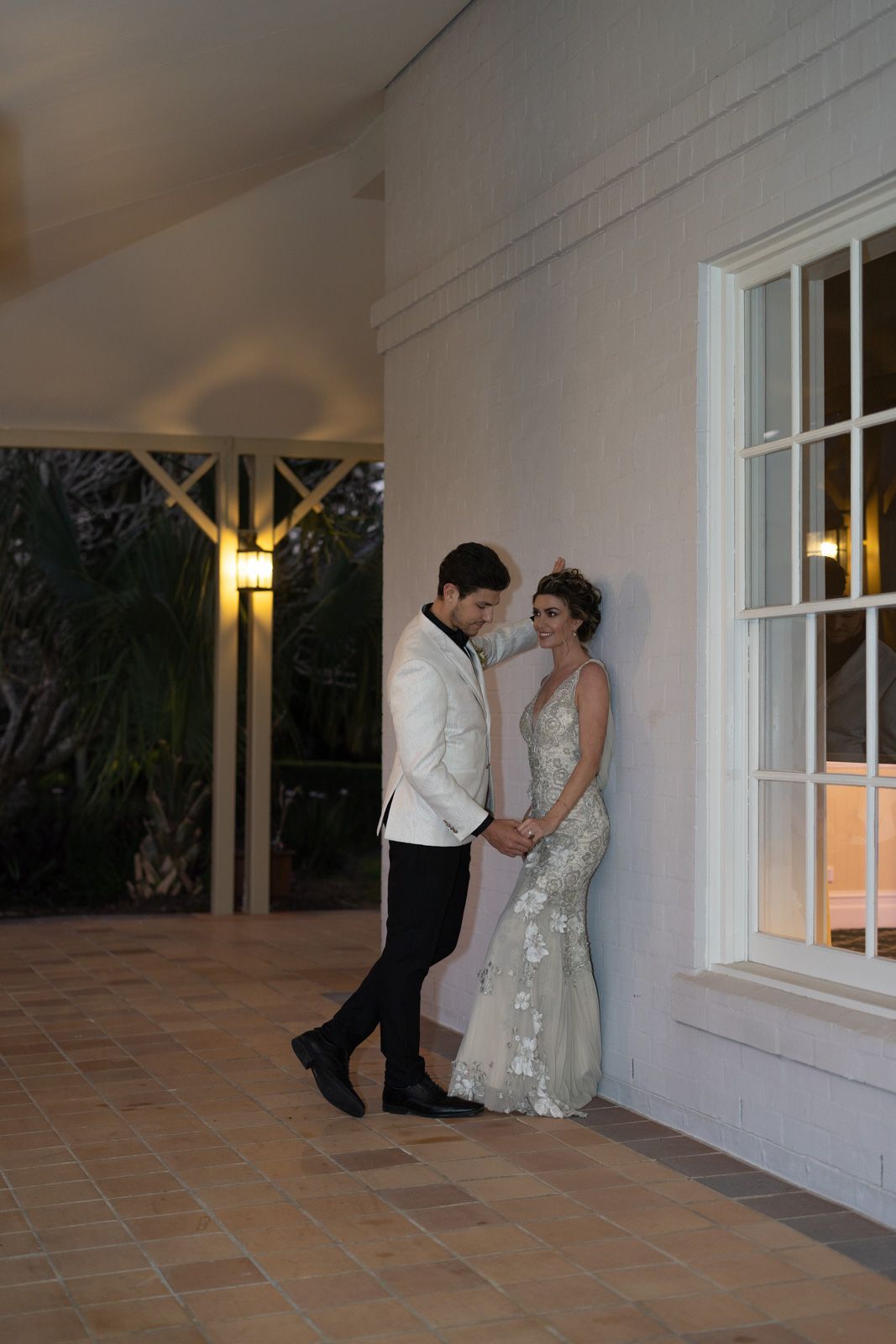 A couple in formal wedding attire share intimate moments against a white wall with windows at dusk.