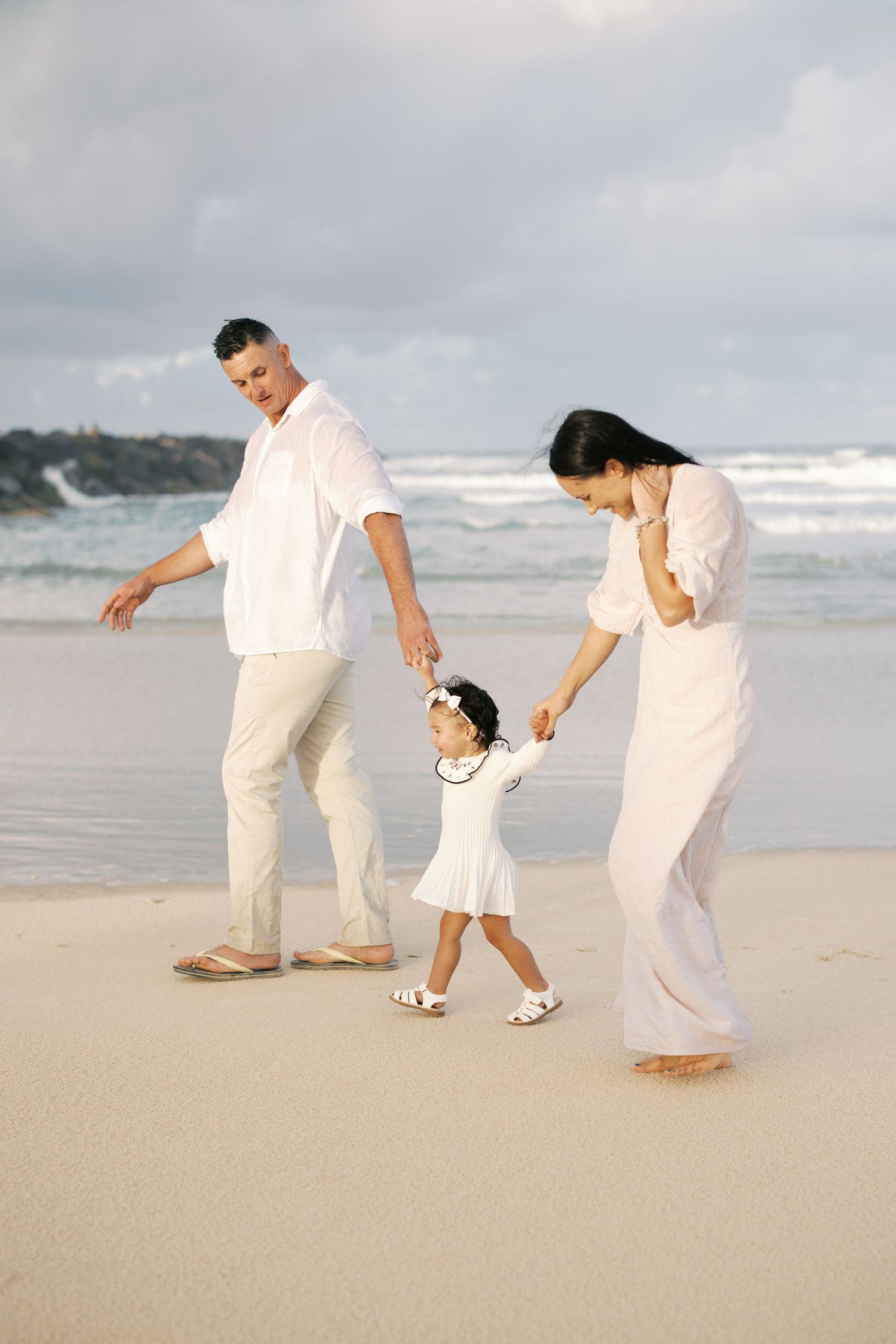 Family enjoying a beach walk together in white clothing against ocean backdrop.