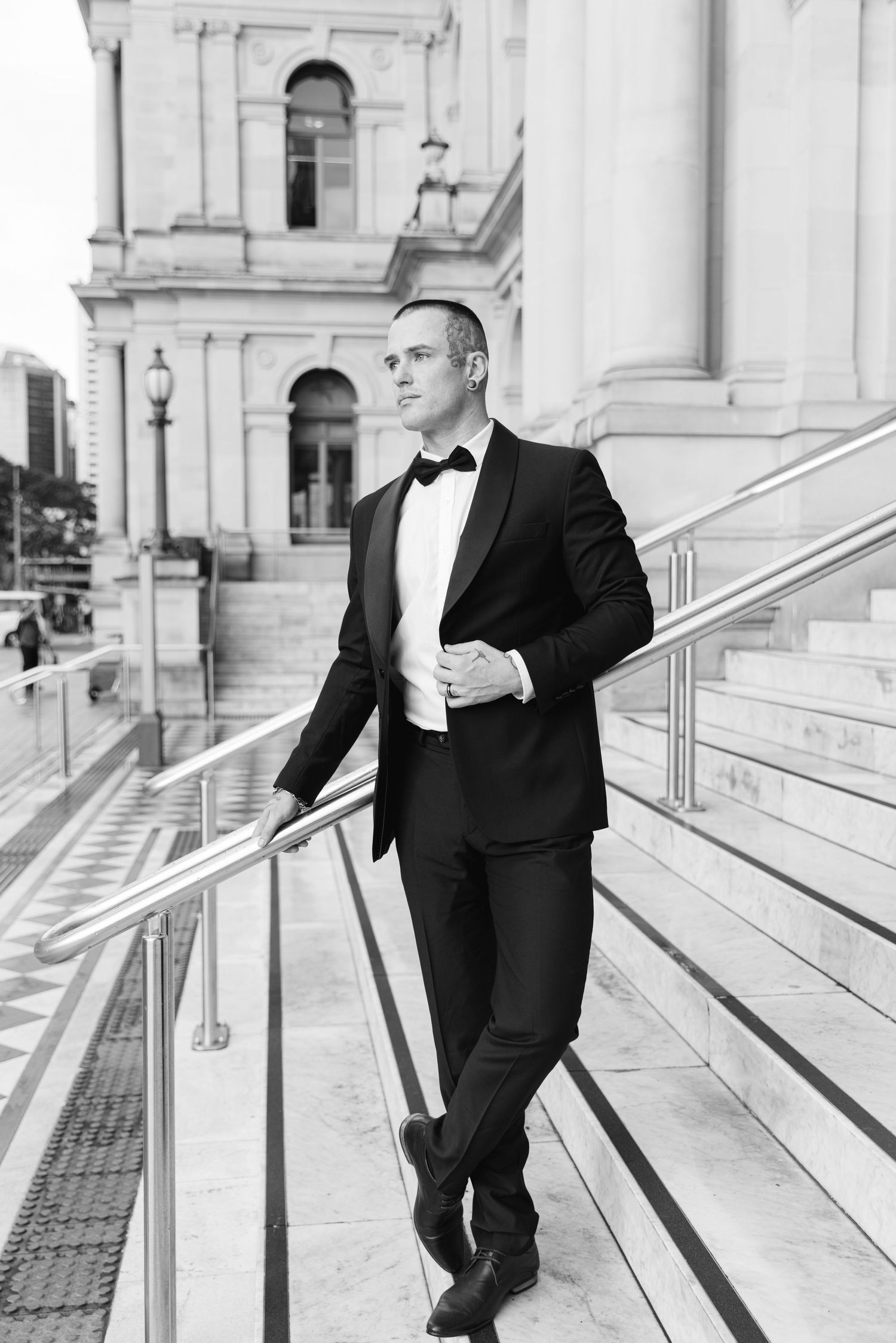 Black and white photo of a person in a classic tuxedo posing on elegant marble steps of a grand building.