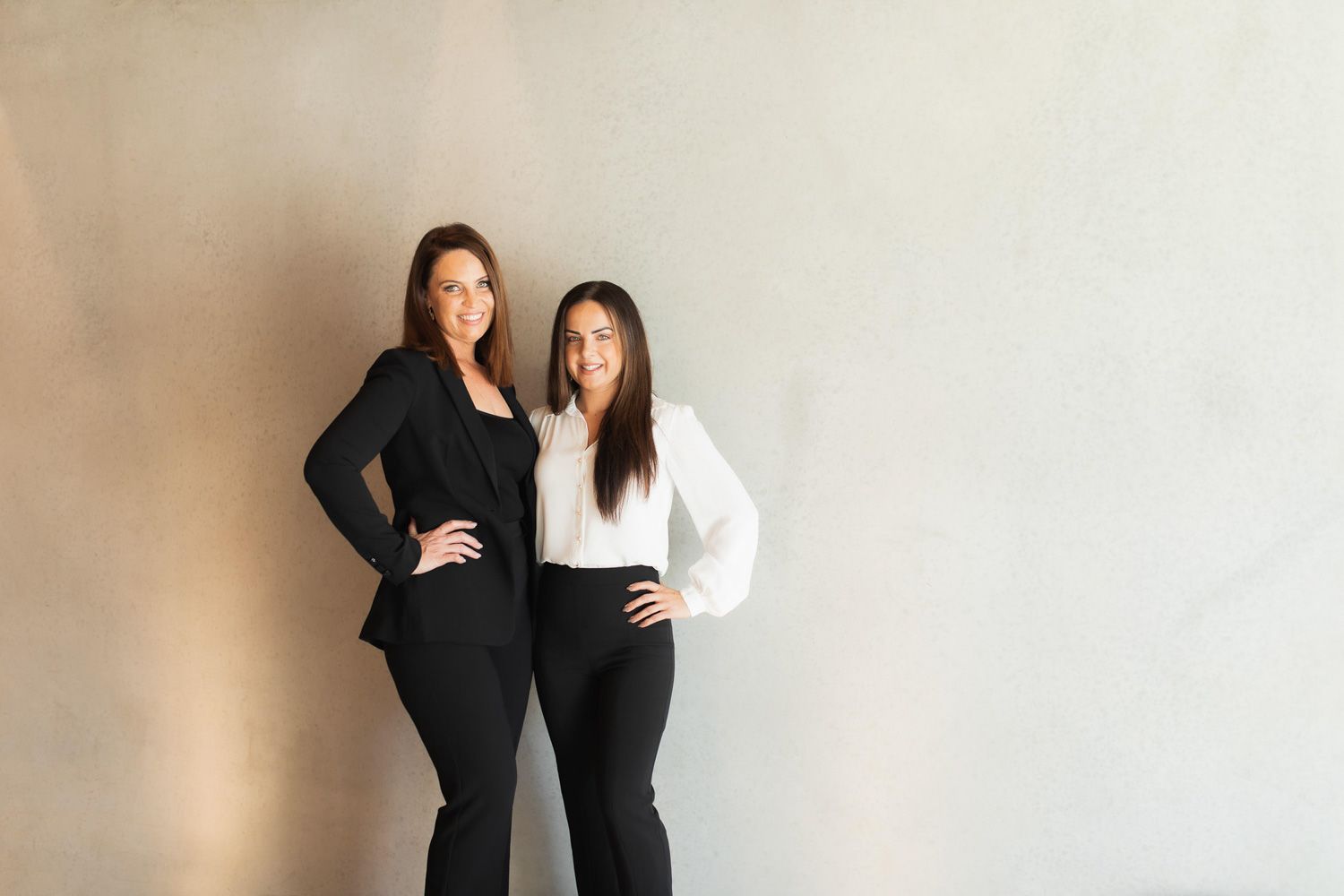 Two business professionals in black and white outfits pose confidently against a neutral wall.