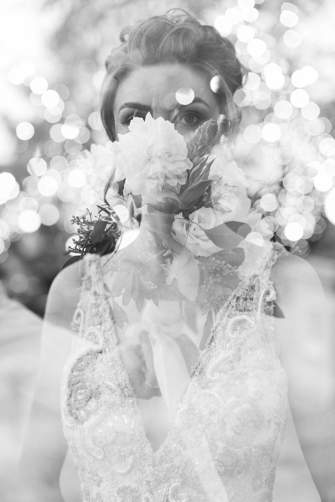 Artistic black and white wedding photo showing a bride holding a bouquet in front of her face with dreamy bokeh effects.