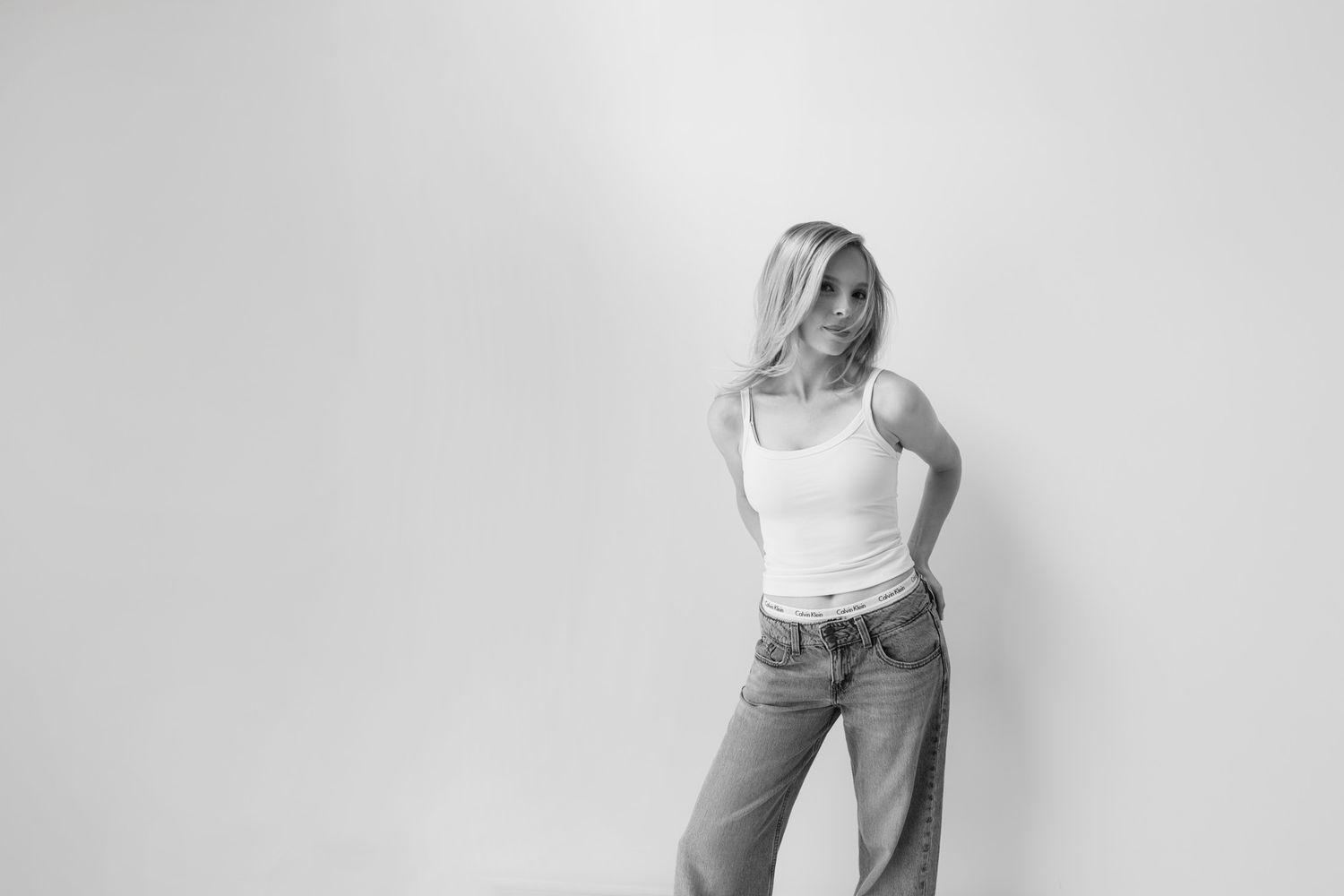 Black and white photo of a blonde woman in a white tank top and jeans posing against a light background.