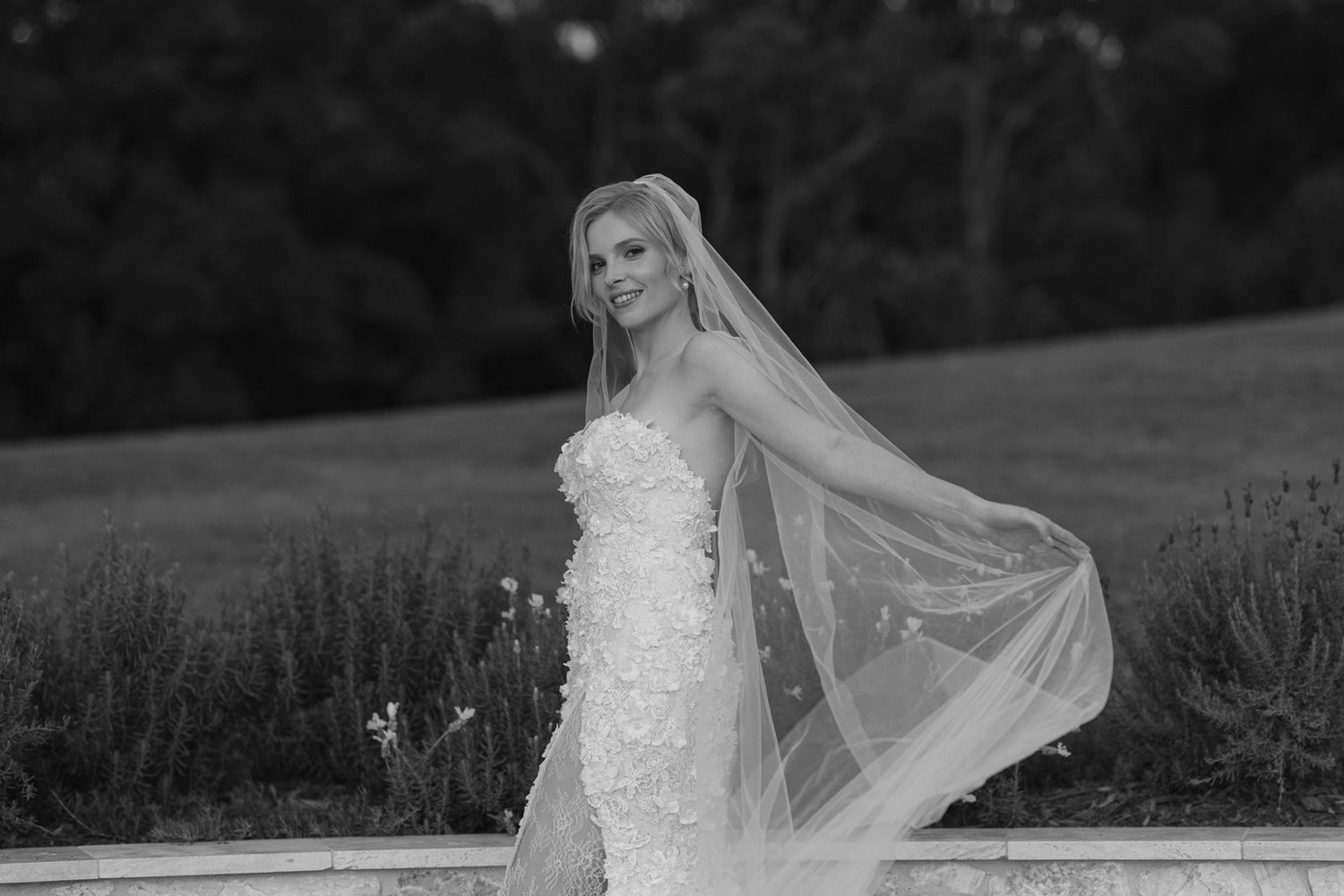 A bride in a flowing white wedding gown and veil poses outdoors in a scenic field during sunset.