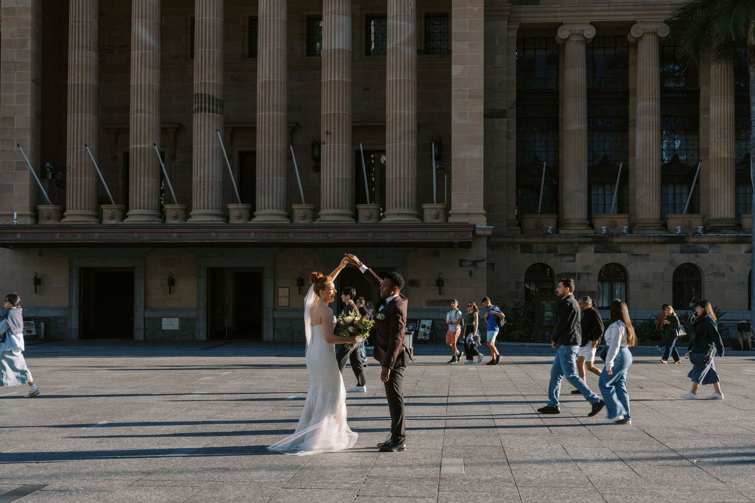 A couple in wedding attire shares a romantic moment in front of a grand columned building while people walk by.