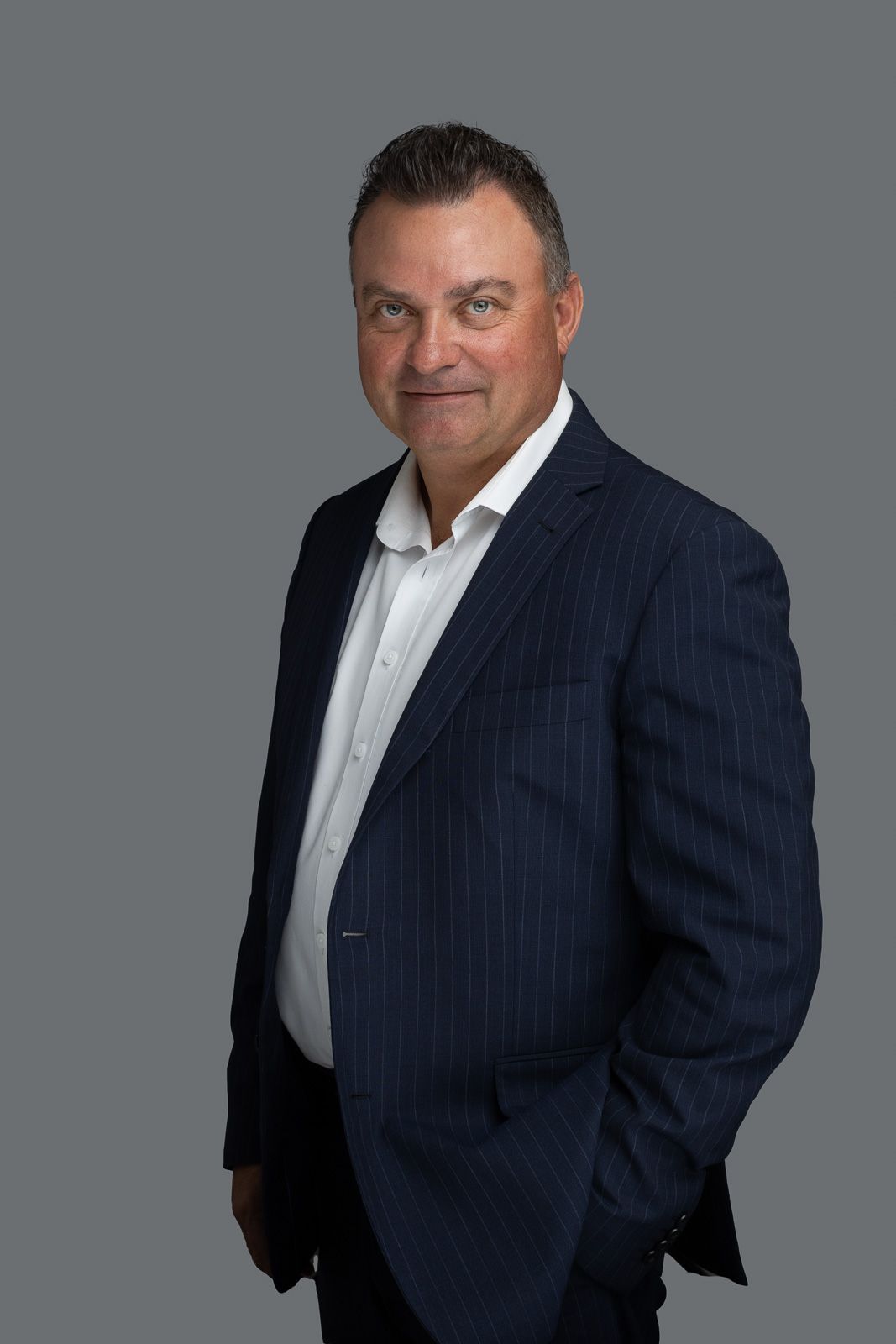Professional headshot portrait of person in navy pinstripe suit jacket and white dress shirt against gray background.