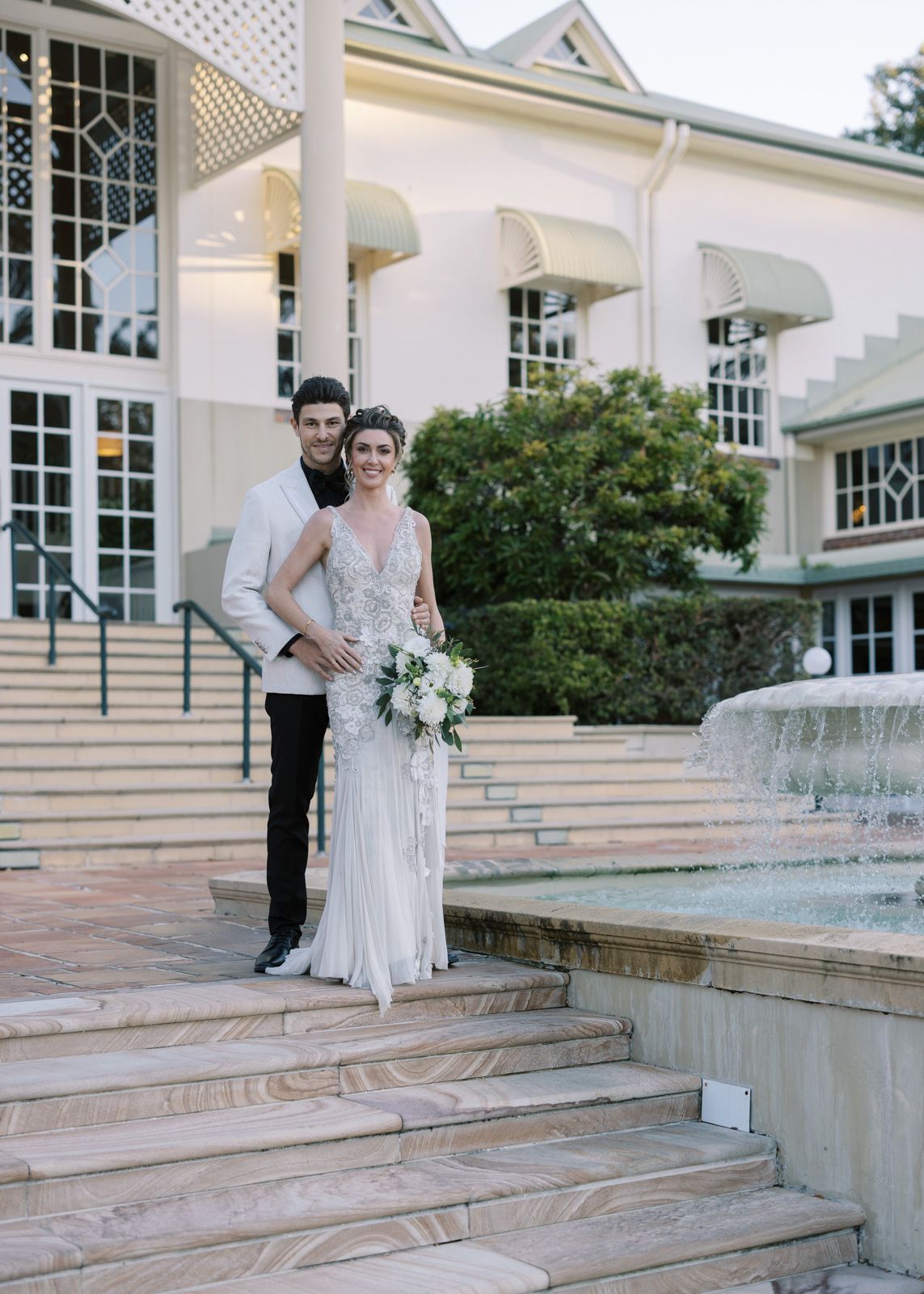 A bride in a white lace gown stands on elegant stone steps outside a white colonial-style building with large windows.