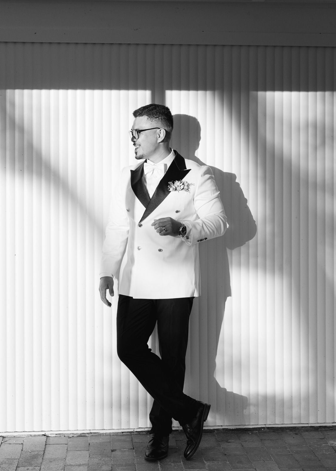 A stylish black and white portrait of someone in a white tuxedo jacket and black pants leaning against a striped wall.
