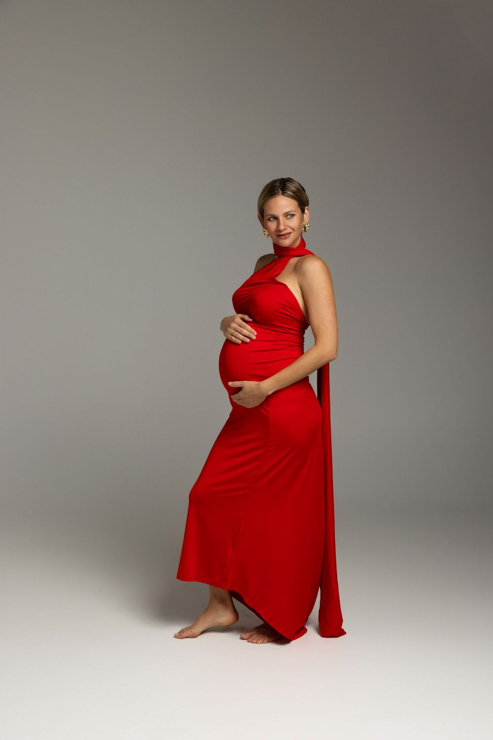 Pregnant woman in elegant red halter dress cradling baby bump, smiling gracefully against gray studio background.
