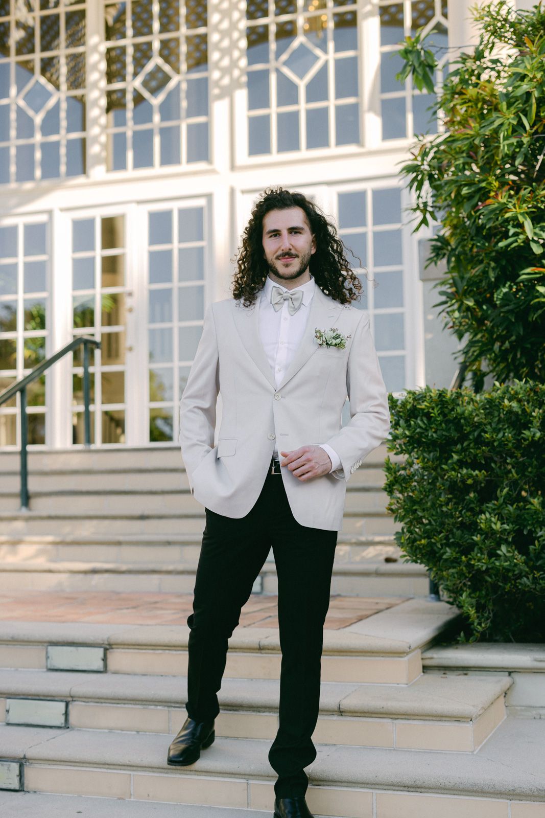 A groom in a light gray blazer and dark pants poses on outdoor steps with decorative windows in the background.