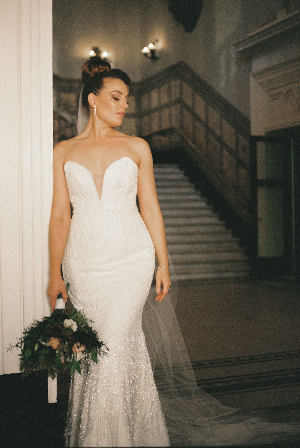 Bride in a strapless beaded mermaid wedding gown with veil holding a bouquet by a grand staircase.